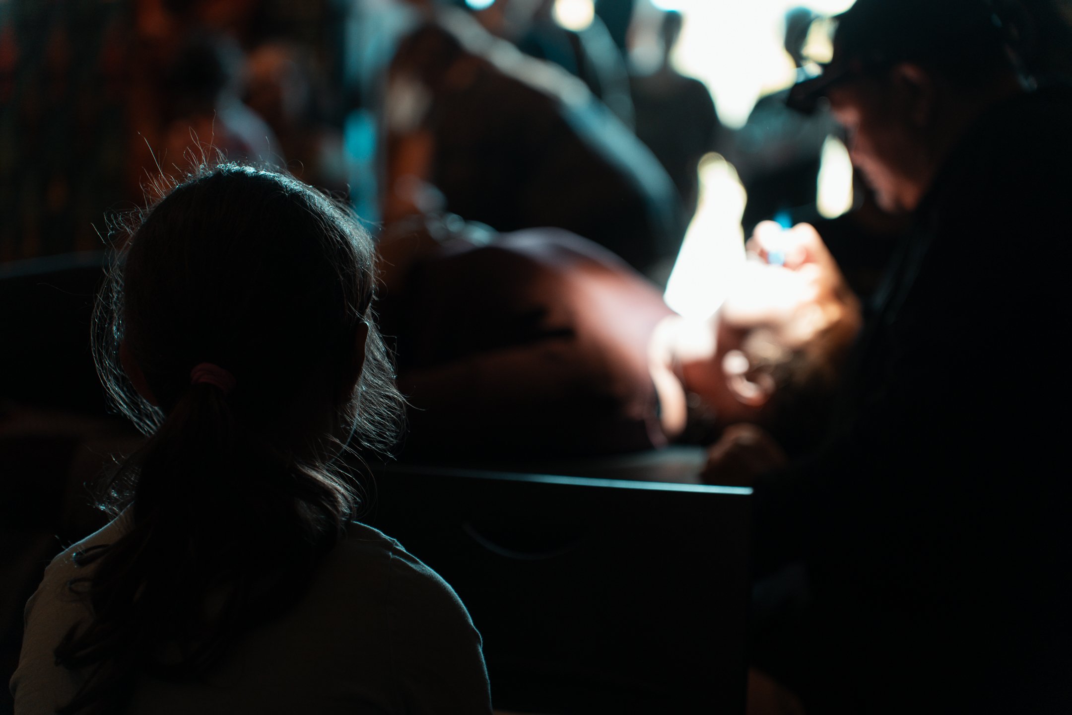 A girl with pulled-back hair watching a medical procedure in a dimly lit room, with a nurse or medical professional attending to a patient.