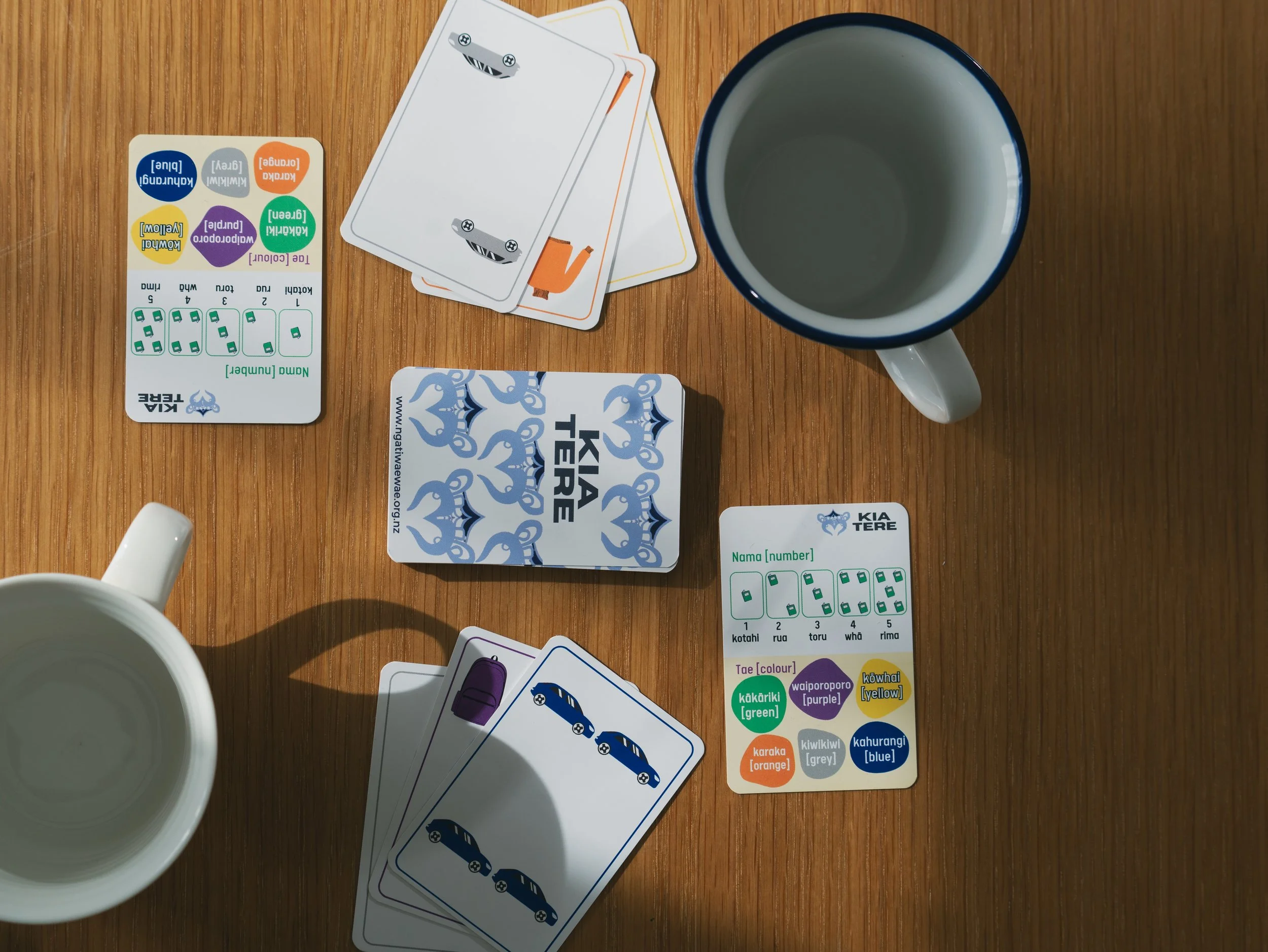 Top-down view of a wooden table with two white coffee mugs, one empty, and several playing cards and colorful game cards.