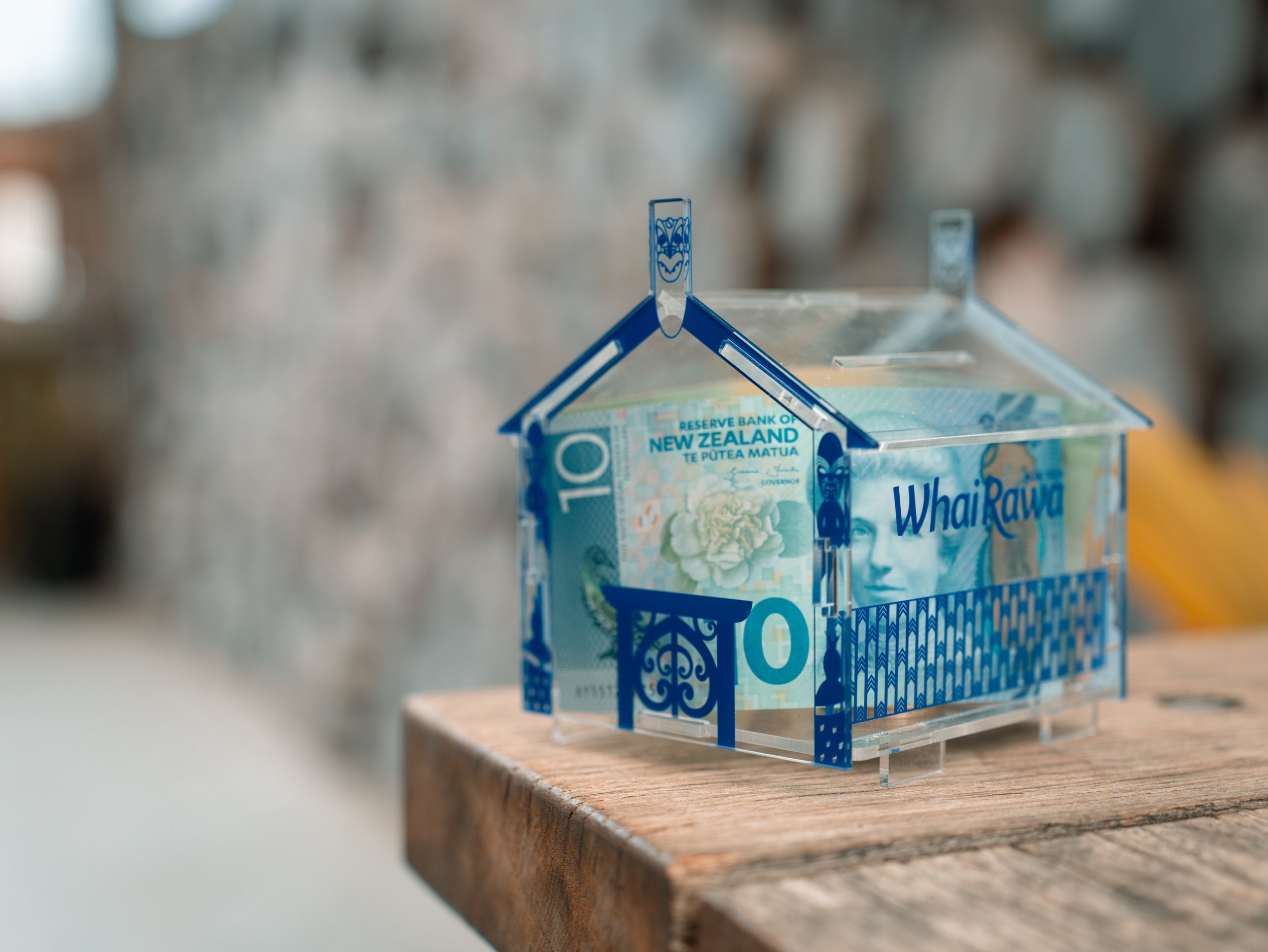 A transparent house-shaped container holds a ten-dollar New Zealand banknote and is placed on a wooden surface.