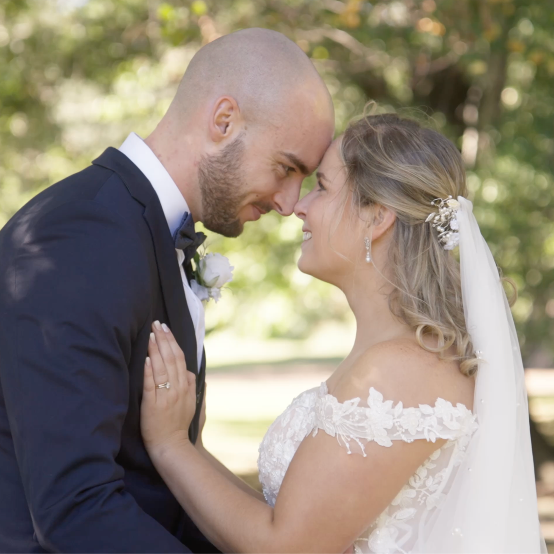A newlywed couple touching foreheads outdoors, smiling, with a blurred green background.