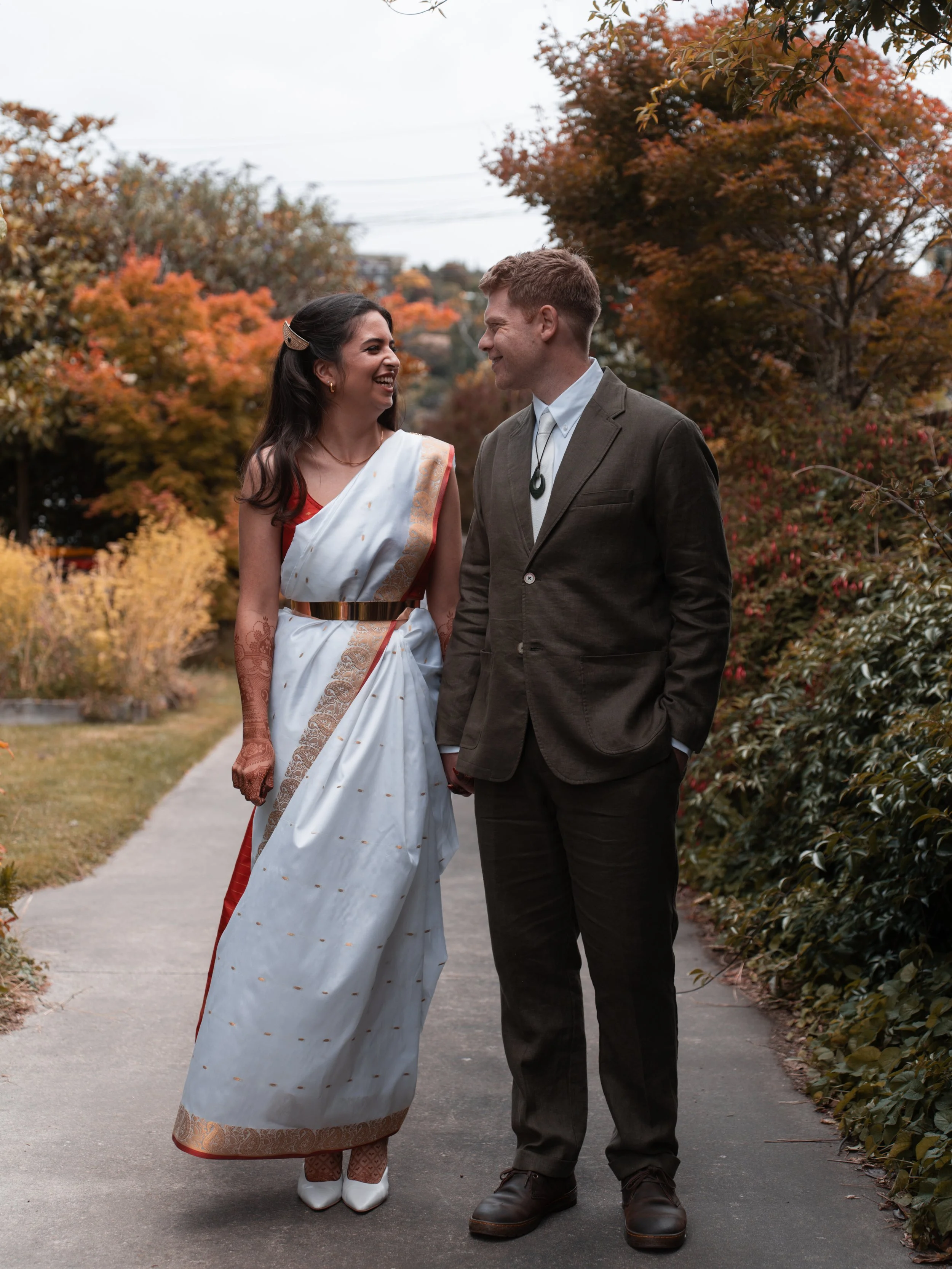 A woman and man are standing on a sidewalk holding hands and smiling at each other, with fall foliage in the background.