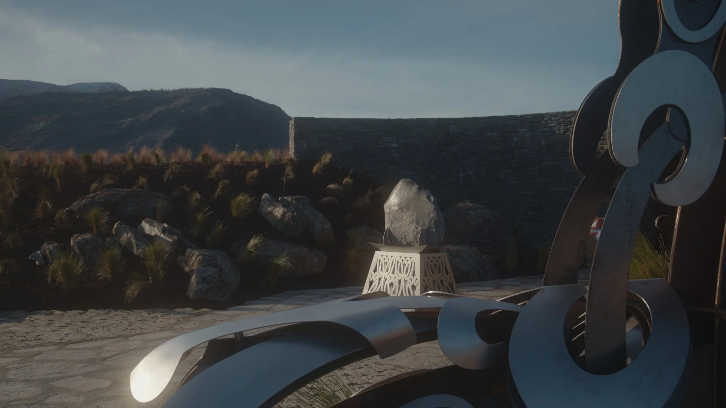 Outdoor sculpture art piece with abstract metal shapes, rocks, and a decorative table, set against a mountainous landscape with a stone wall and cloudy sky.