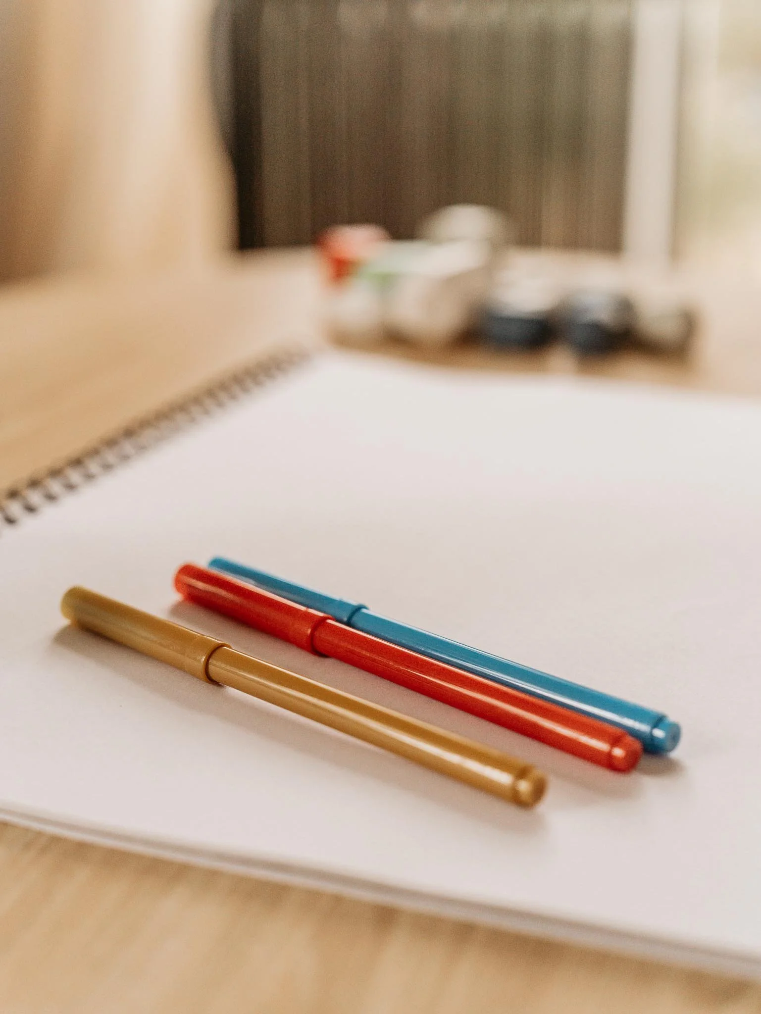 Three colorful pens (gold, red, blue) placed on a white notebook on a wooden surface with blurred objects in the background.