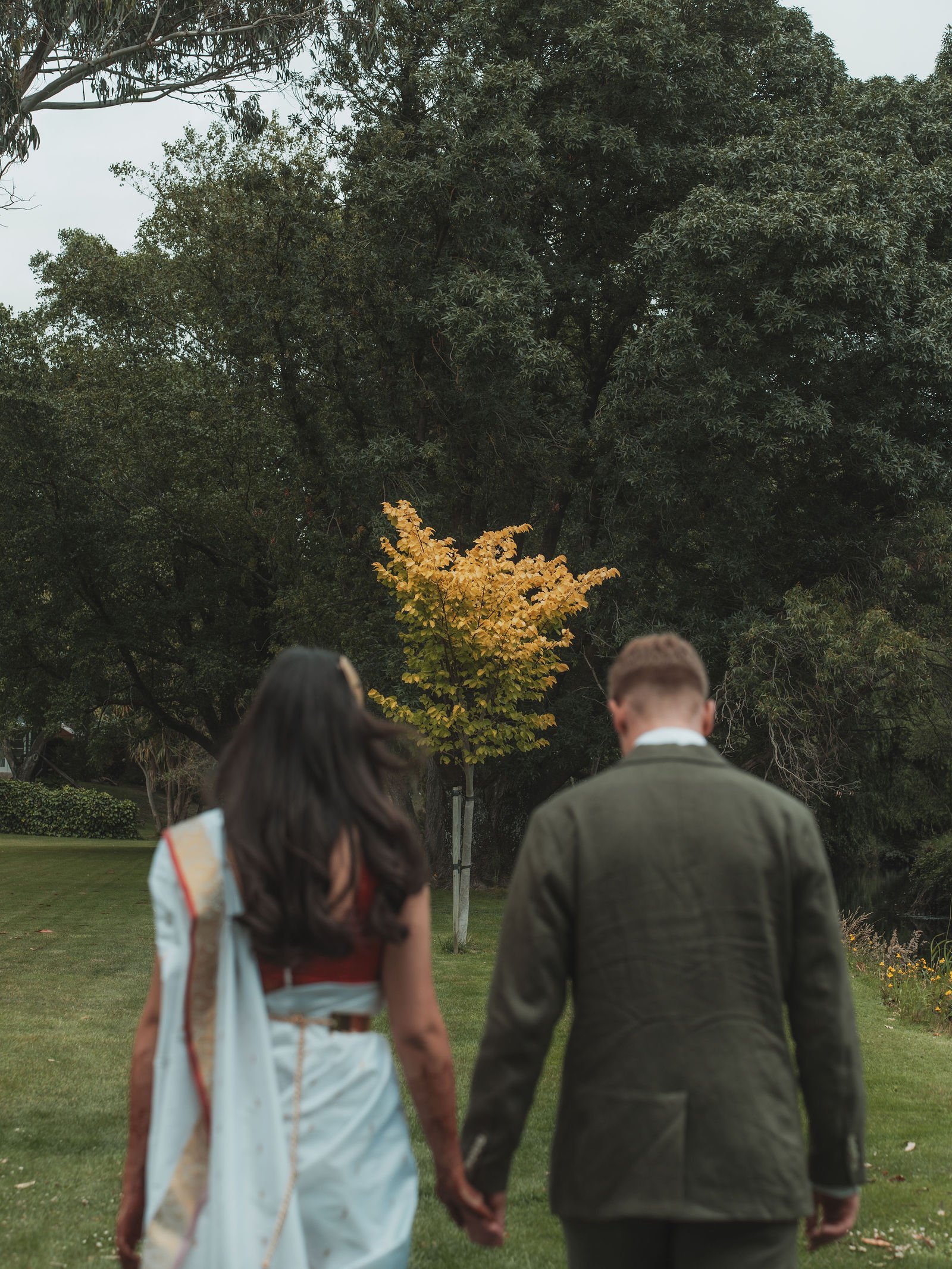 A young woman and man walking hand in hand on a park pathway with green grass and trees in the background.