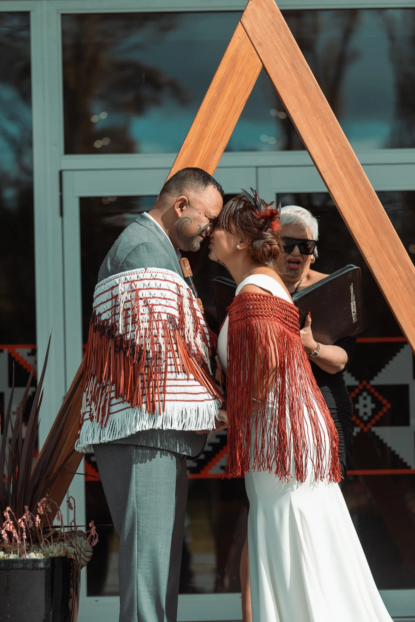 A couple getting married touching foreheads under a wooden arch, with a officiant in the background.