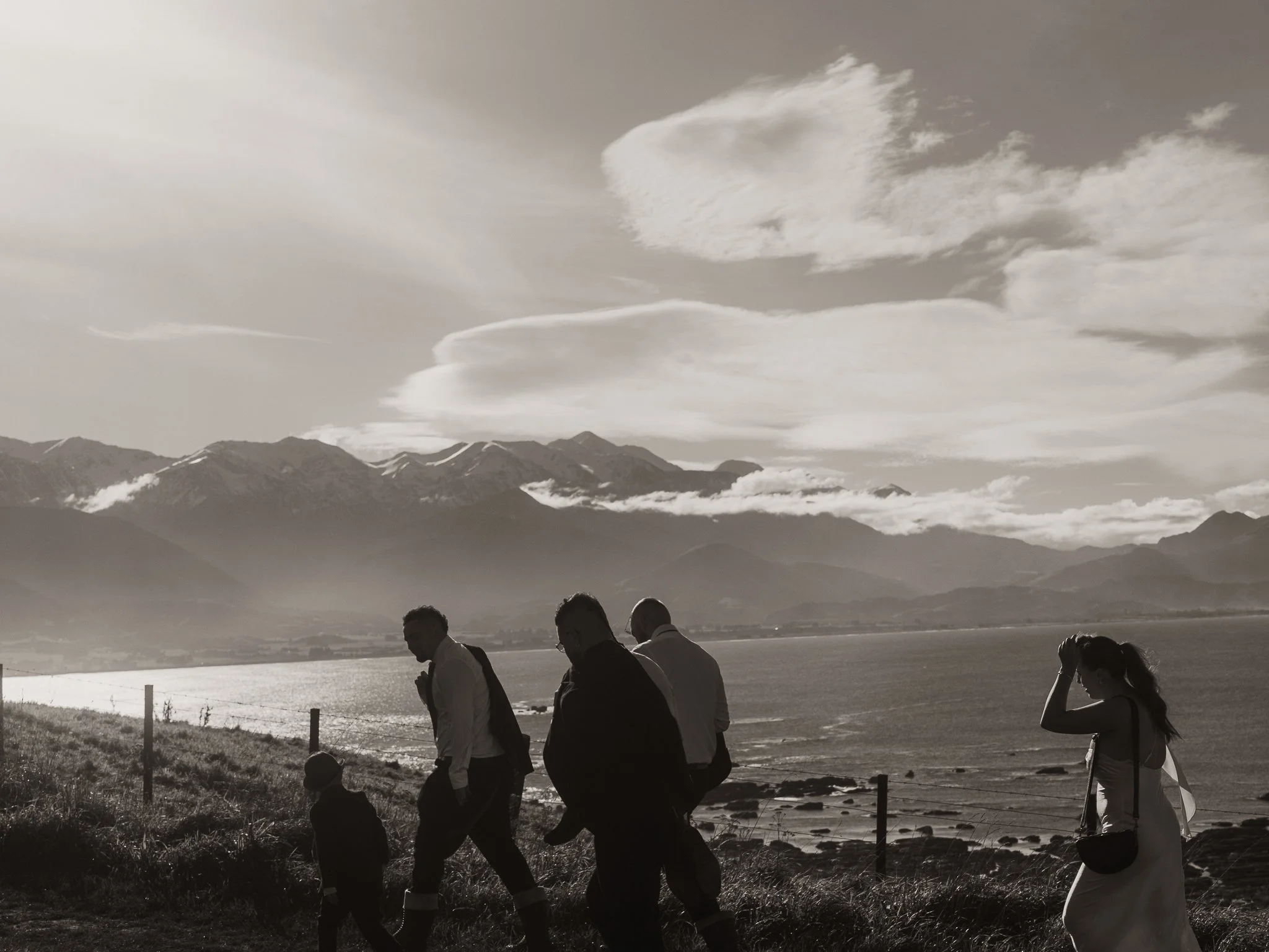 A black and white photo of five people walking along a coastal area with mountains and clouds in the background. One woman appears to be adjusting her hair or head.