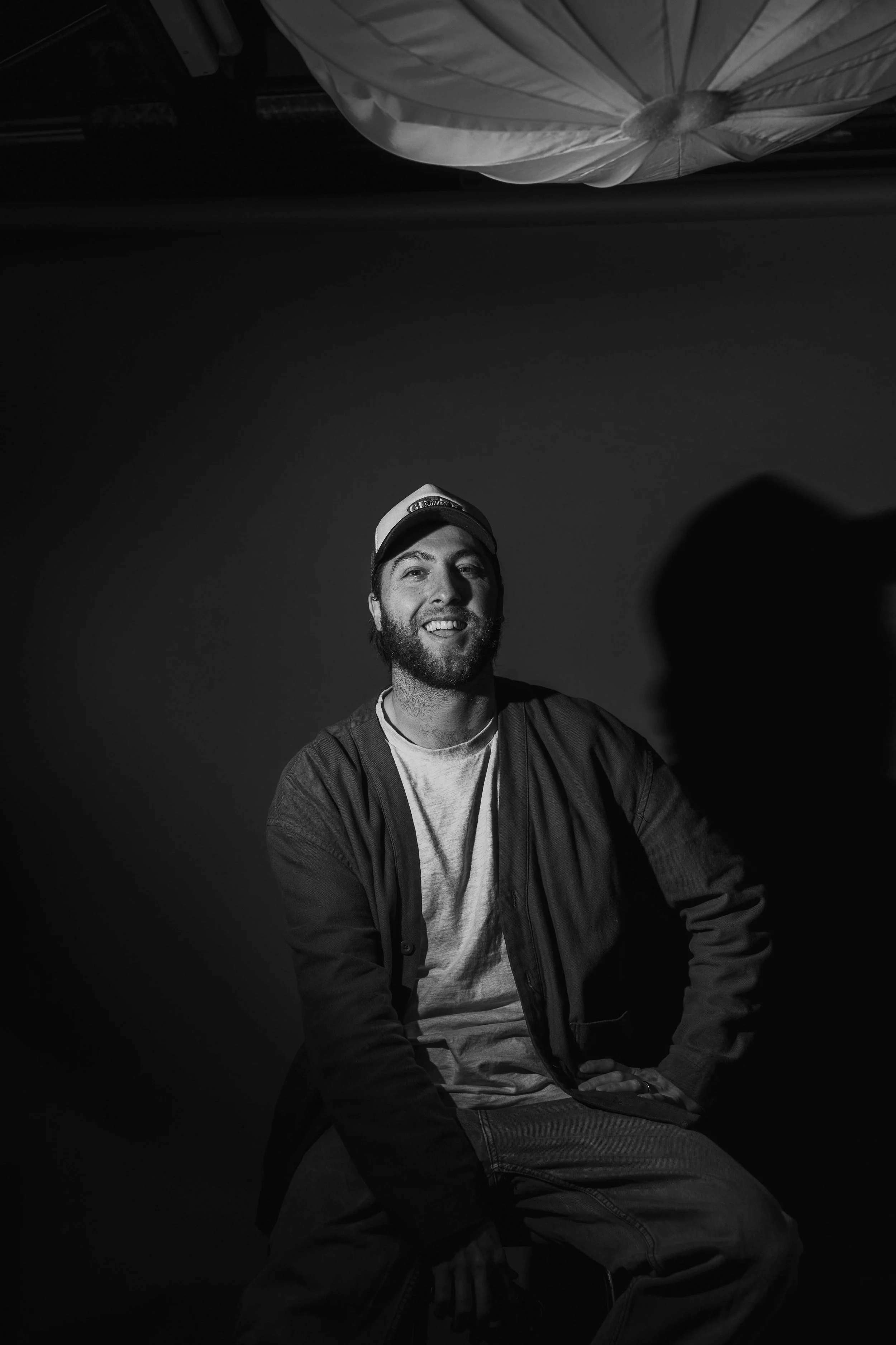 Black and white photo of a man with a beard smiling, wearing a baseball cap, a T-shirt, and a jacket, seated against a plain background.