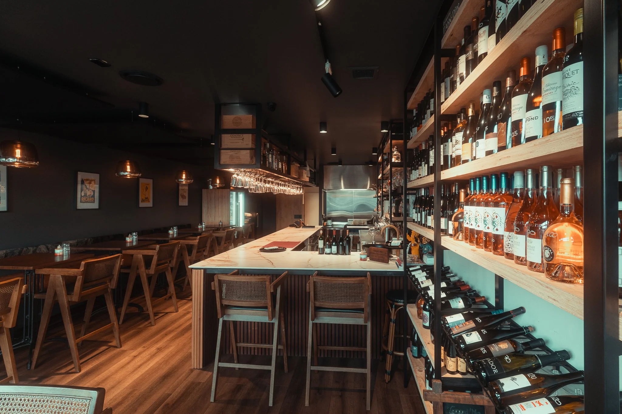 Inside a modern bar with wine bottles displayed on wooden shelves, a marble-topped island, wooden chairs, and tables along a dark wall with framed artwork.