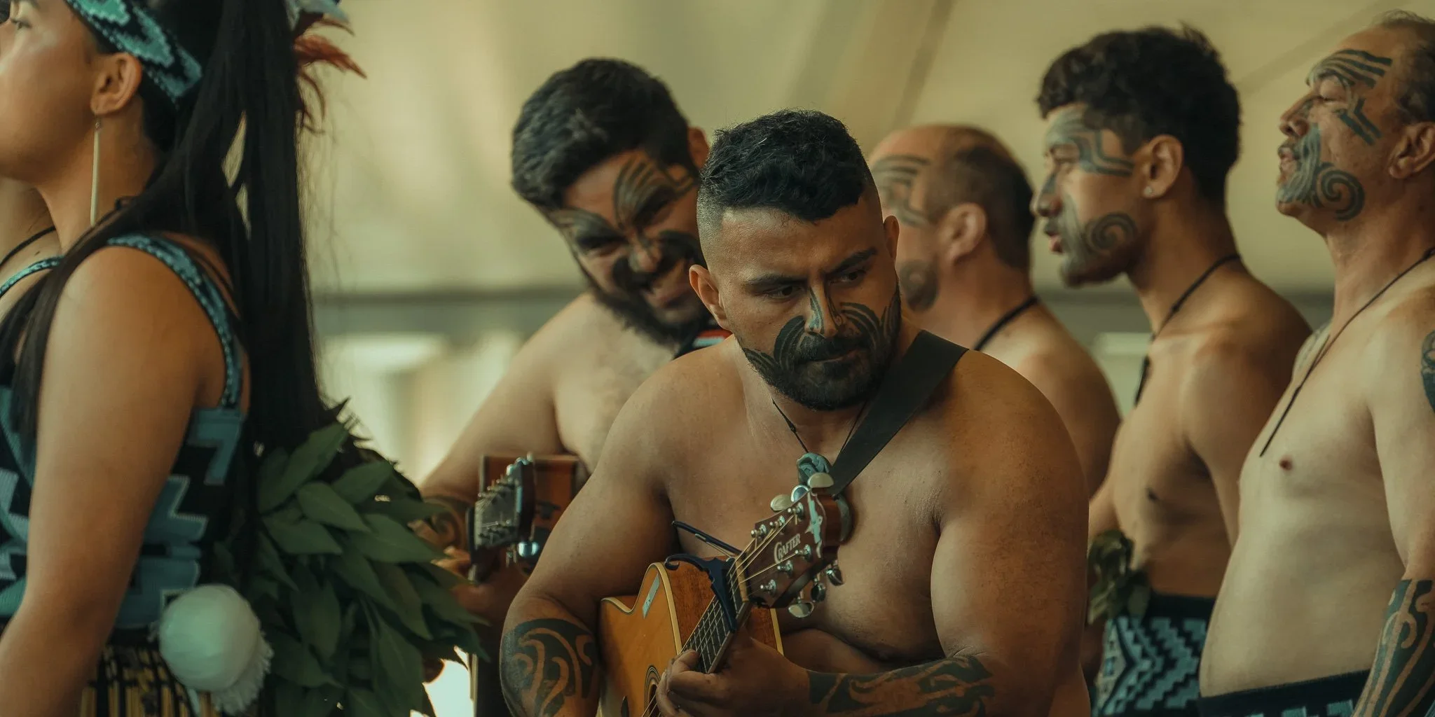 A group of men and women with traditional Maori face tattoos, some shirtless, one playing a guitar, standing and looking serious in a cultural setting.