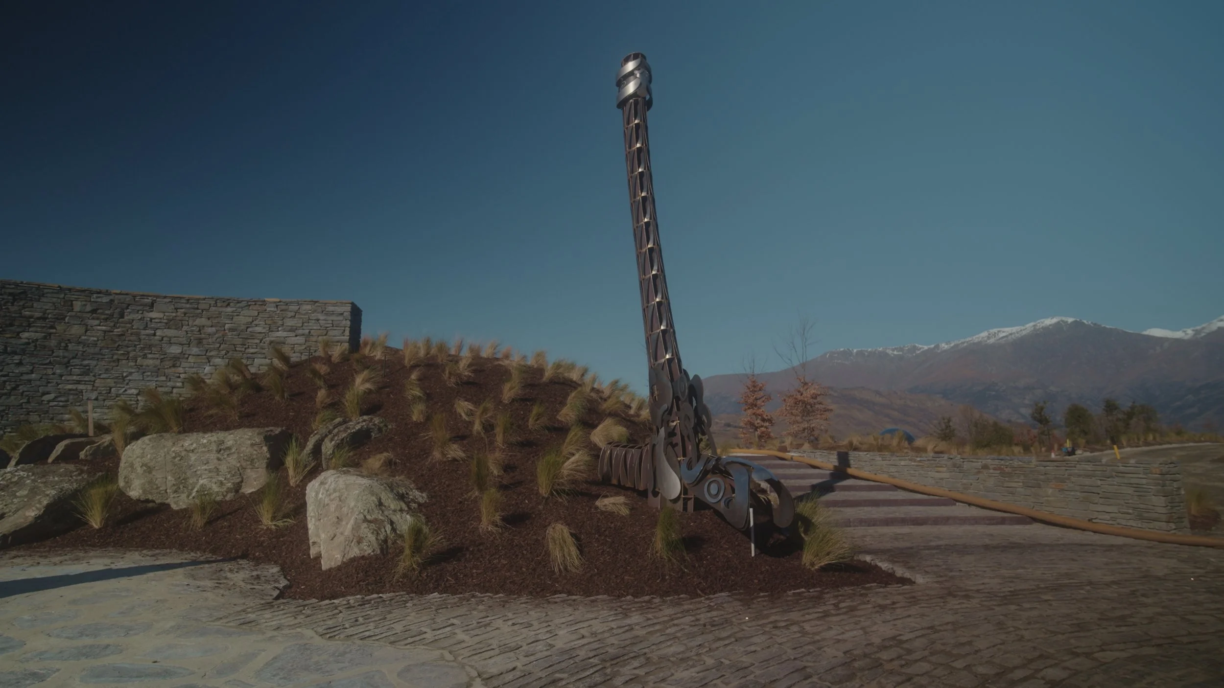 A large metal sculpture resembling a tall, slender giraffe standing on a landscaped hillside with rocks and plants, against a background of mountains and a clear blue sky.