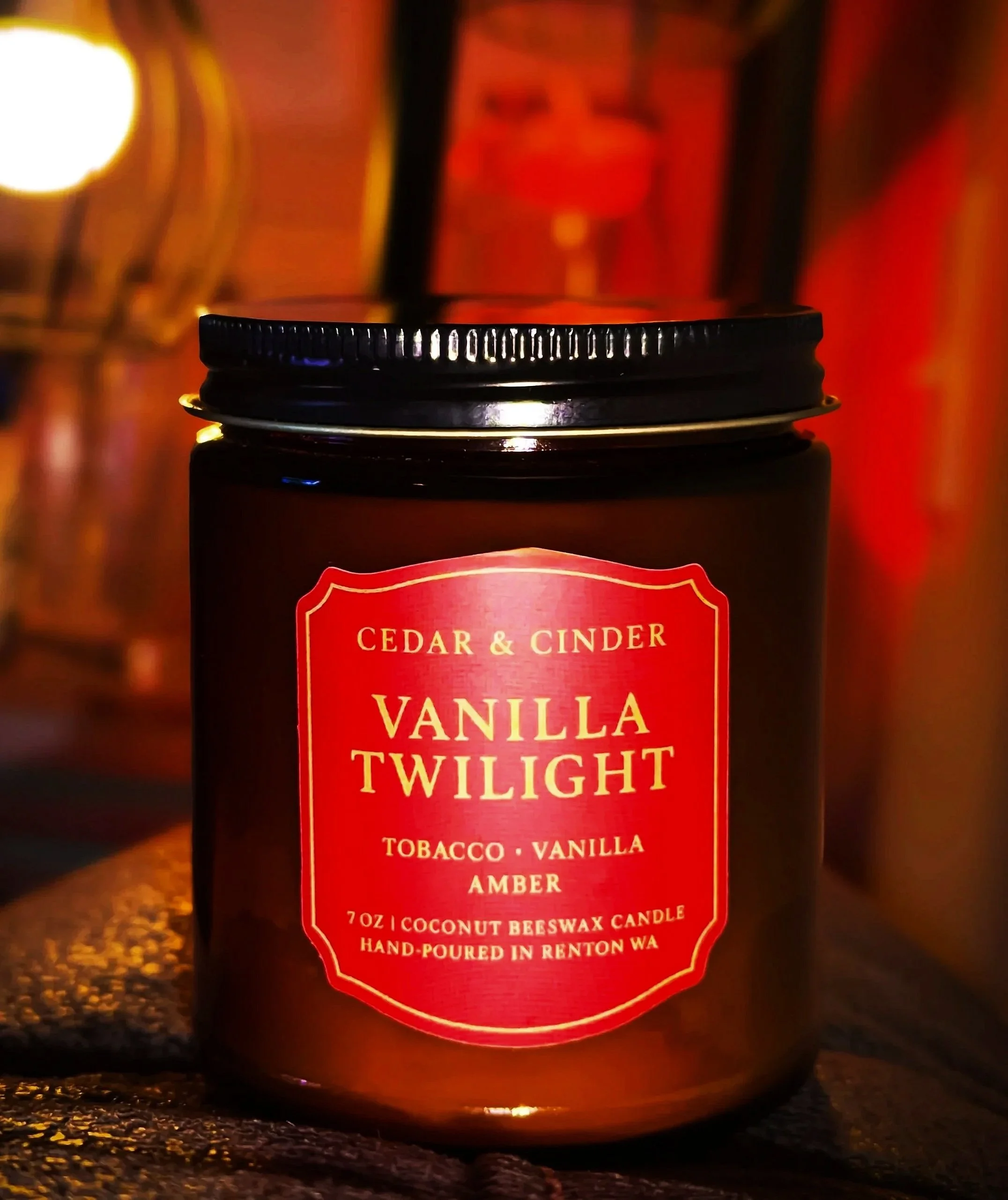 A candle in a dark amber jar with a black lid, labeled "Vanilla Twilight" by Cedar & Cinder. The label states it is a coconut beeswax candle with vanilla and amber scent, in Forks, Washington.