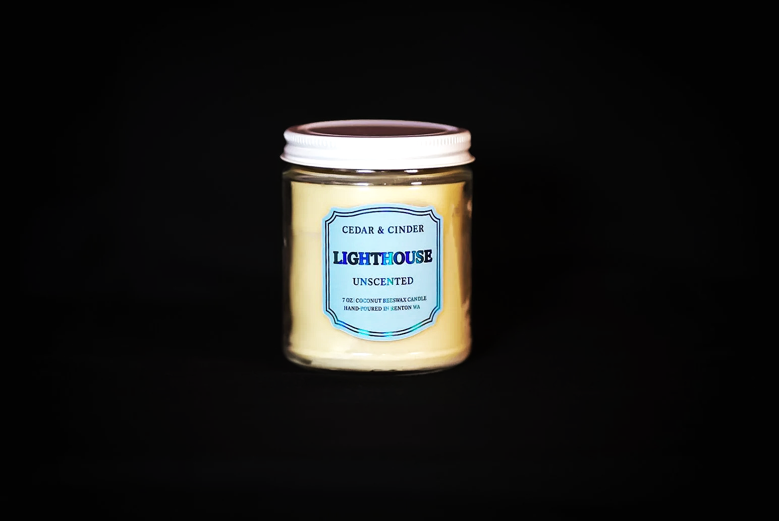 A candle in a glass jar with a white lid, labeled 'Lighthouse,' scented with cedar and cinder, unscented, from Renton, WA.