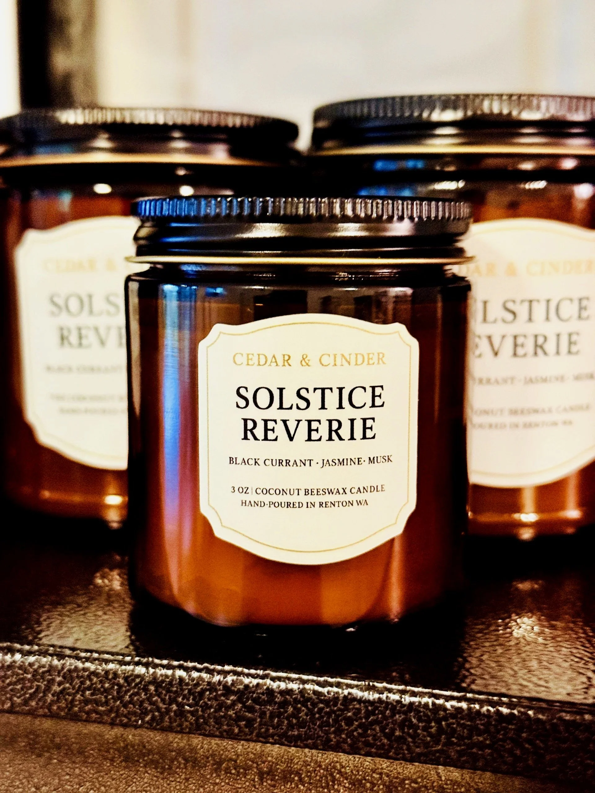 A brown candle jar labeled 'Solstice Reverie' with a white label, featuring scents of black currant, jasmine, and musk, with a black lid, placed on a dark surface. In the background, two more similar candles are visible.