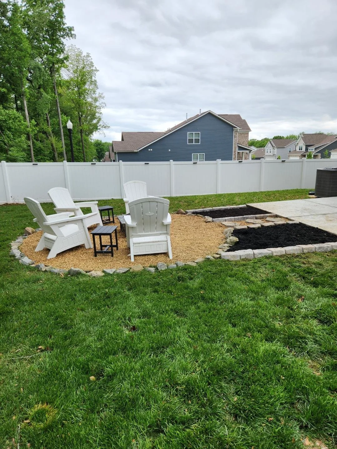 Backyard with white Adirondack chairs and small black tables on a gravel area, surrounded by green lawn, with a white privacy fence, houses in the background, and cloudy sky.
