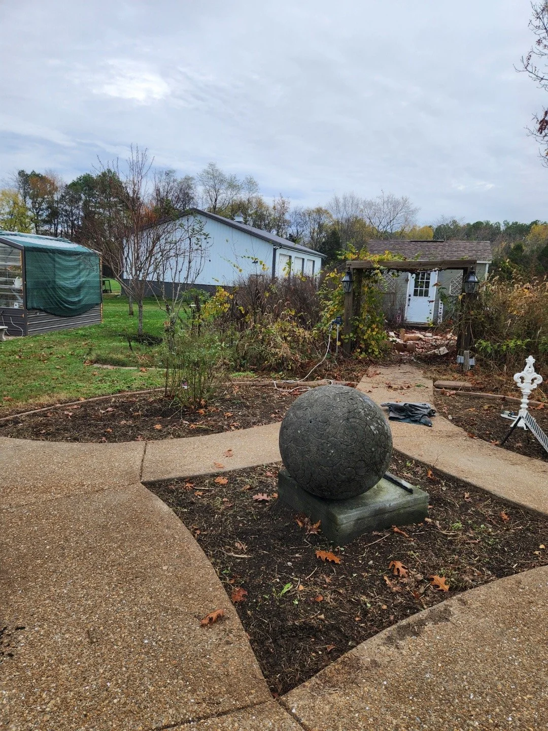 A backyard with a concrete pathway, a spherical stone sculpture, leafless trees, a small garden shed with lights, and a larger white building in the background, under a cloudy sky.