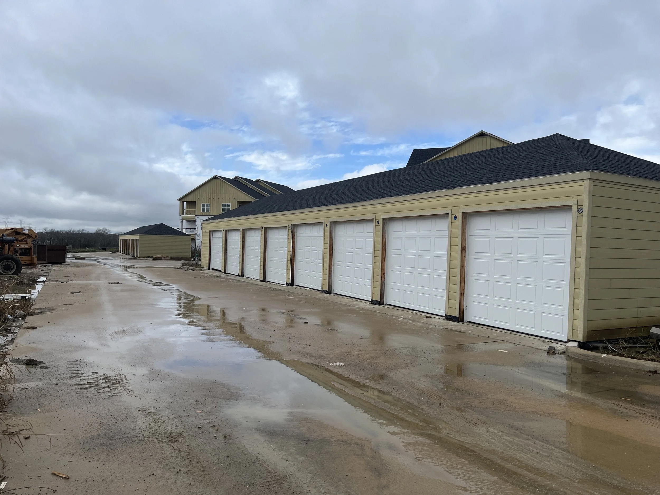 Construction site with newly built garage units with white doors, a gravel driveway with puddles, and houses under construction in the background under cloudy skies.