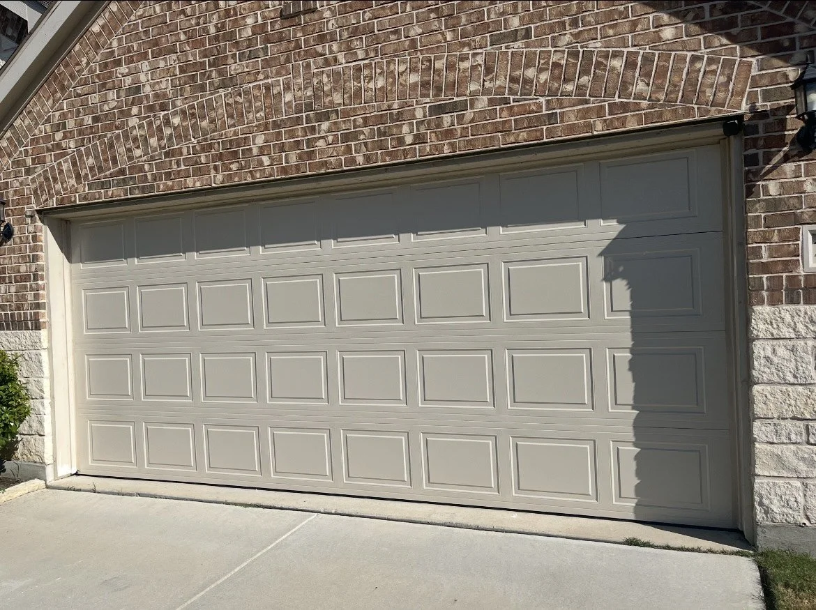 Closed beige sectional garage door in front of brick and stone house with a small black outdoor light fixture on the right.