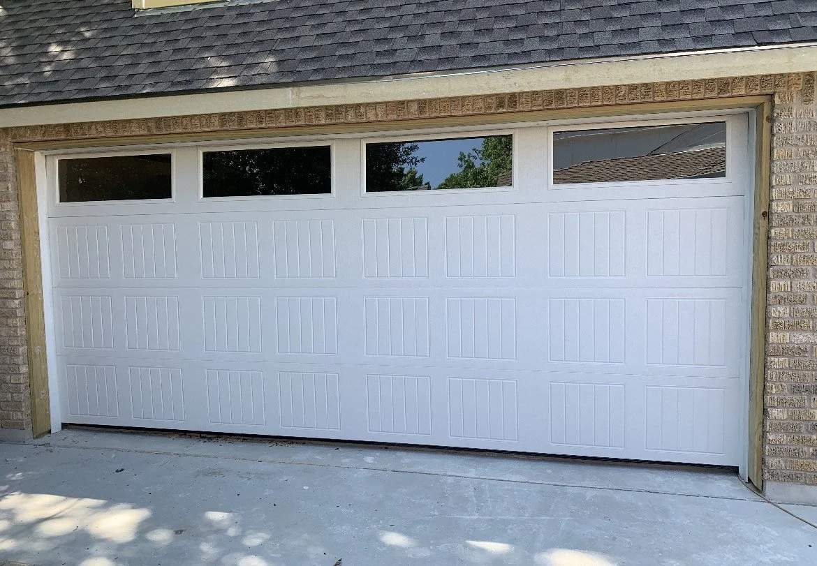 New white garage door with four windows at the top, set in a brick house with a shingled roof.