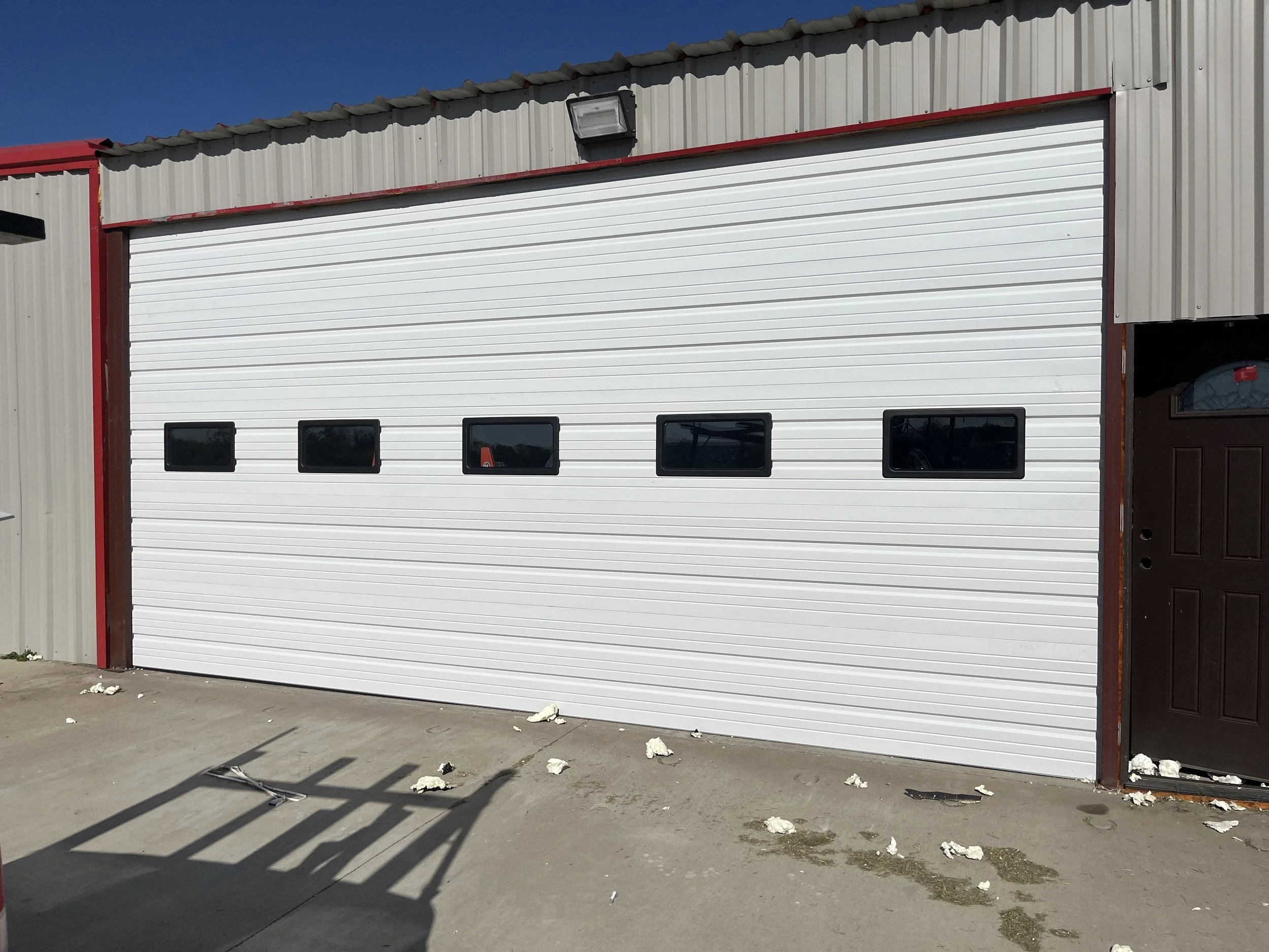 White industrial garage door with four horizontal windows, mounted on a building with beige metal siding, red trim, and a brown door to the right.