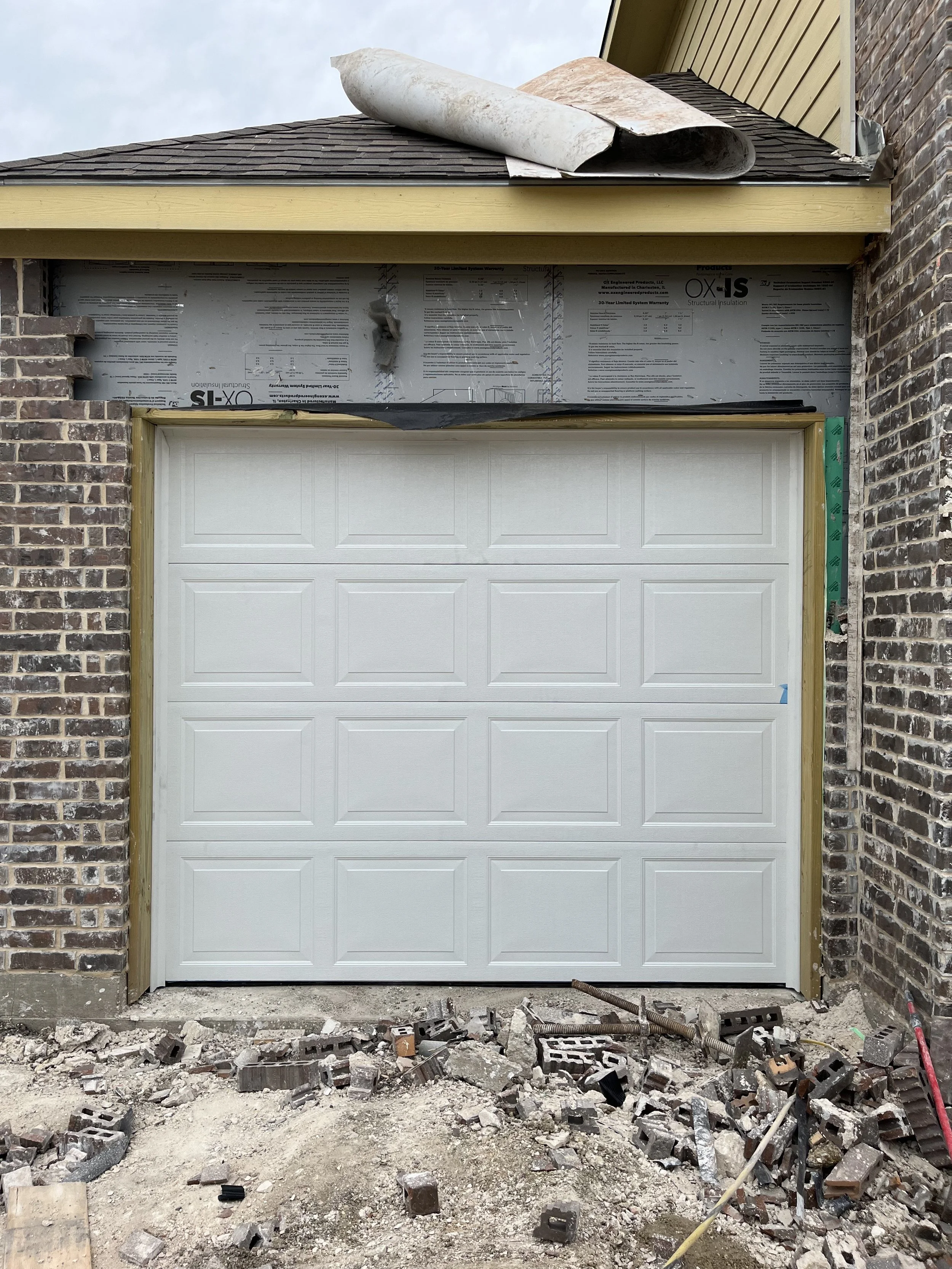 Garage door installation under construction with bricks and debris on the ground and roof work in progress.