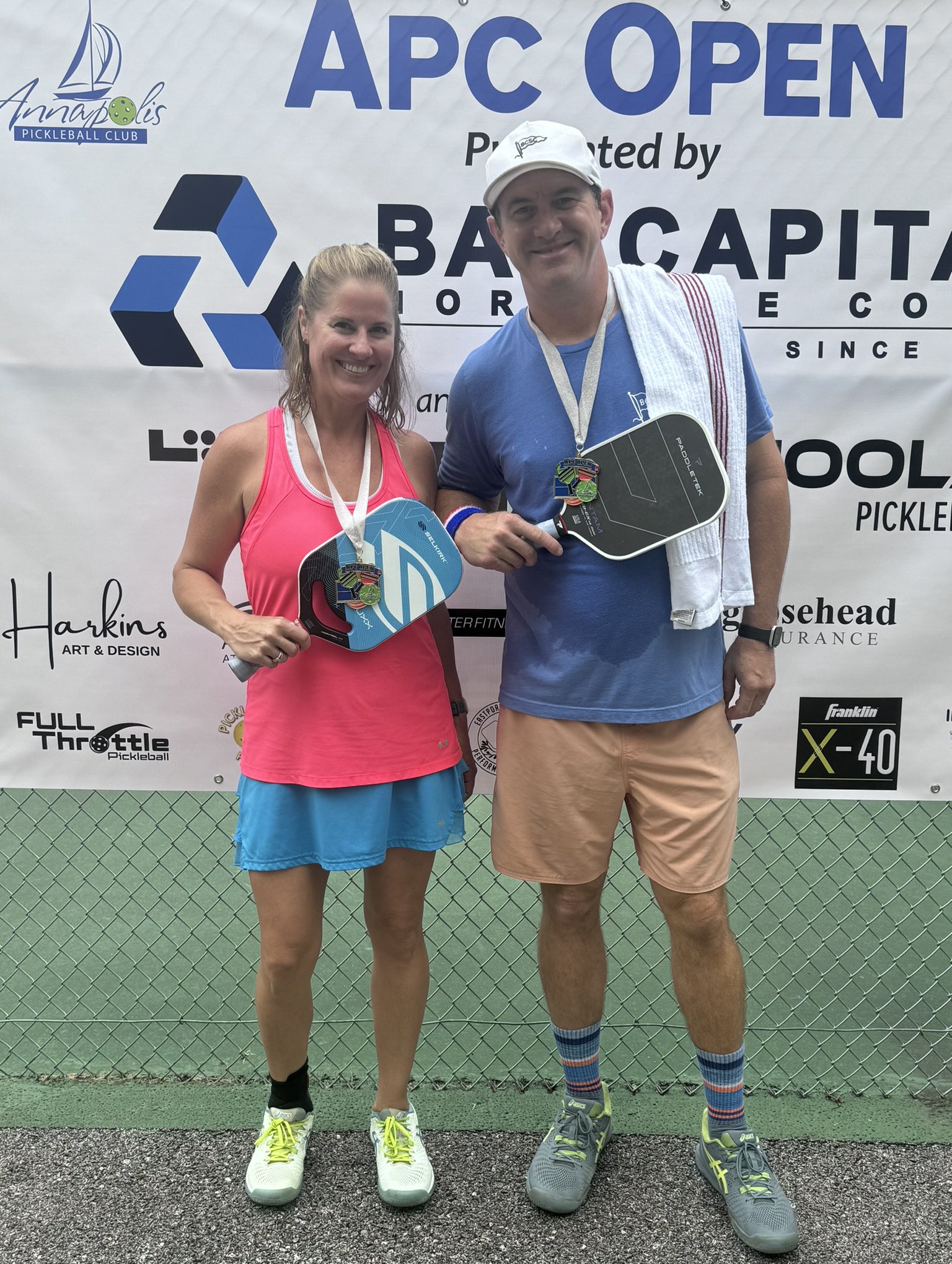 Two satisfied pickleball players, a woman in a pink tank top and blue shorts and a man in a blue t-shirt and peach shorts, holding paddles and medals, standing in front of a banner that reads 'APC OPEN'.