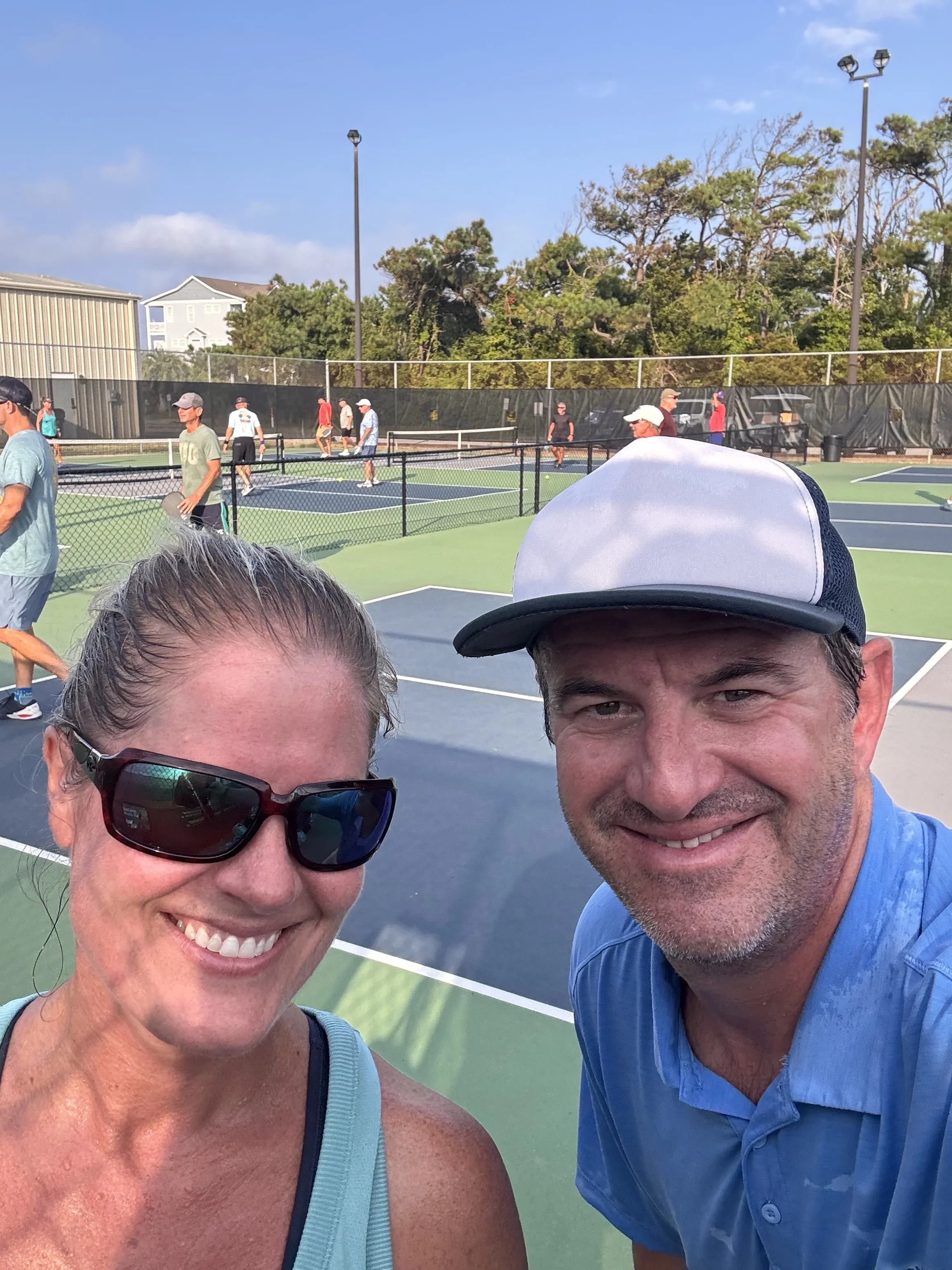Smiling man and woman taking a selfie at a tennis court, with several people playing tennis in the background on a sunny day.