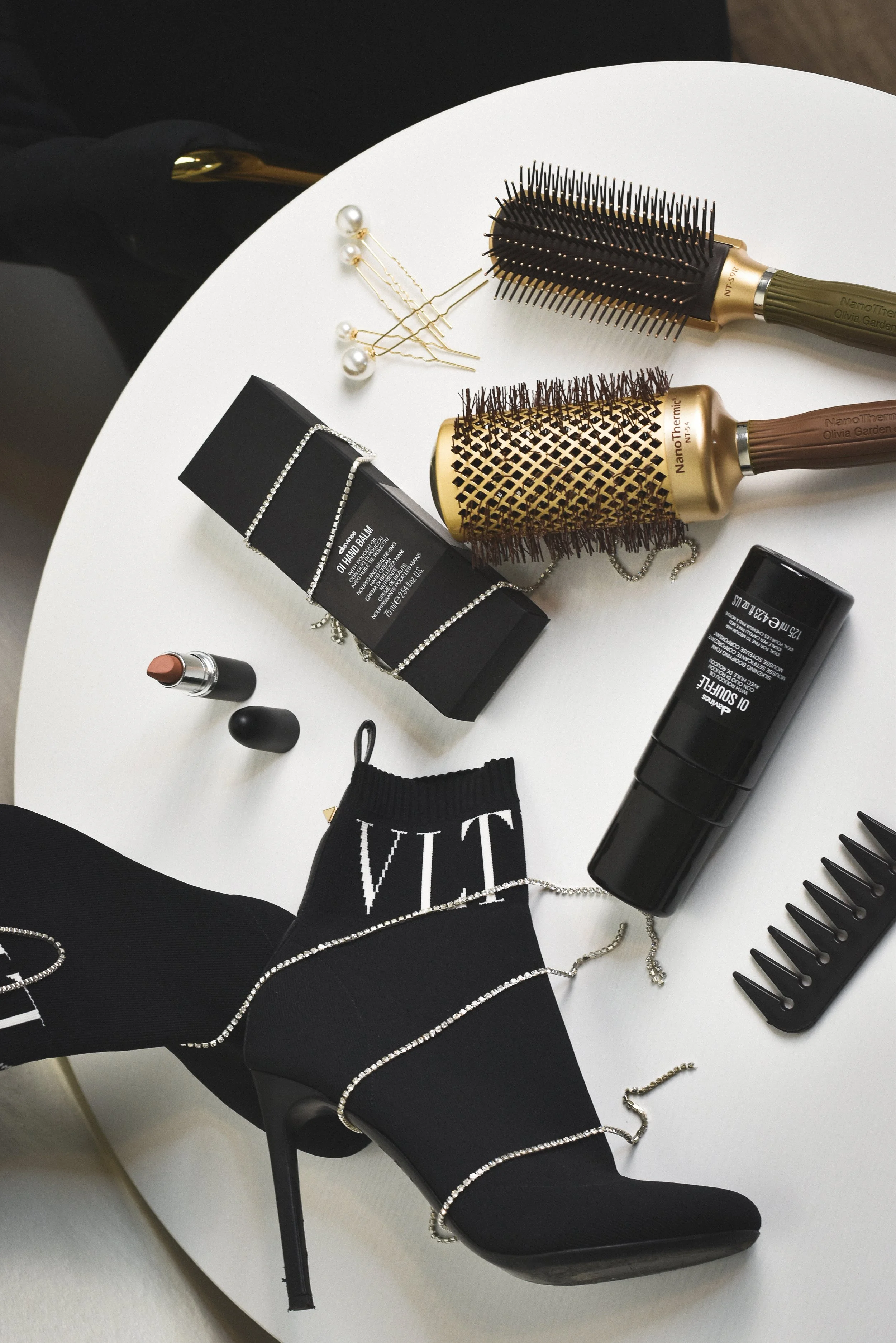 A white table with a black high-heeled sock shoe, jewelry, hairbrushes, hairpins, lipstick, and skincare products on top.