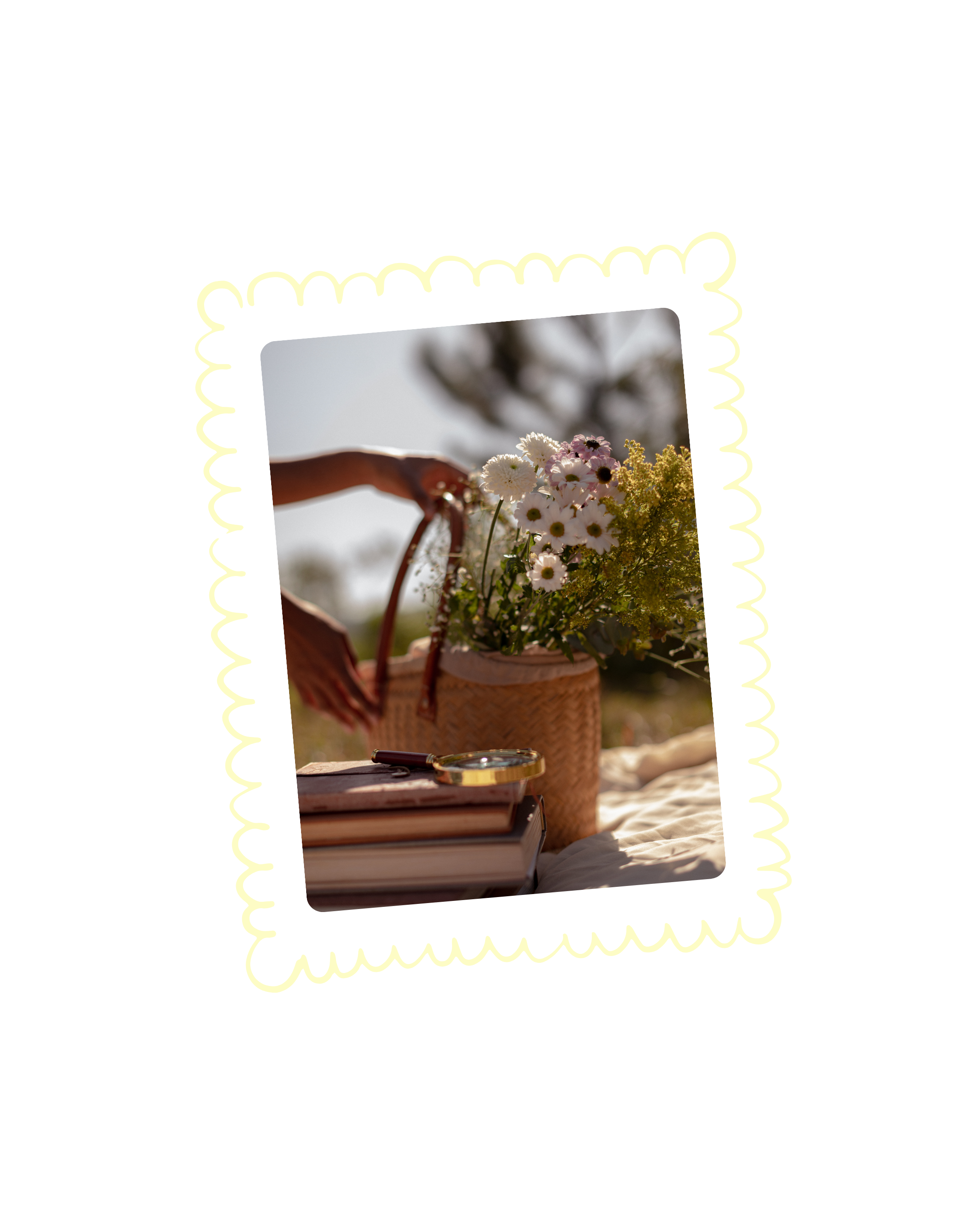 A picnic setup on a blanket with a wicker basket filled with white daisies and other flowers, a stack of books, and a magnifying glass in warm natural sunlight.