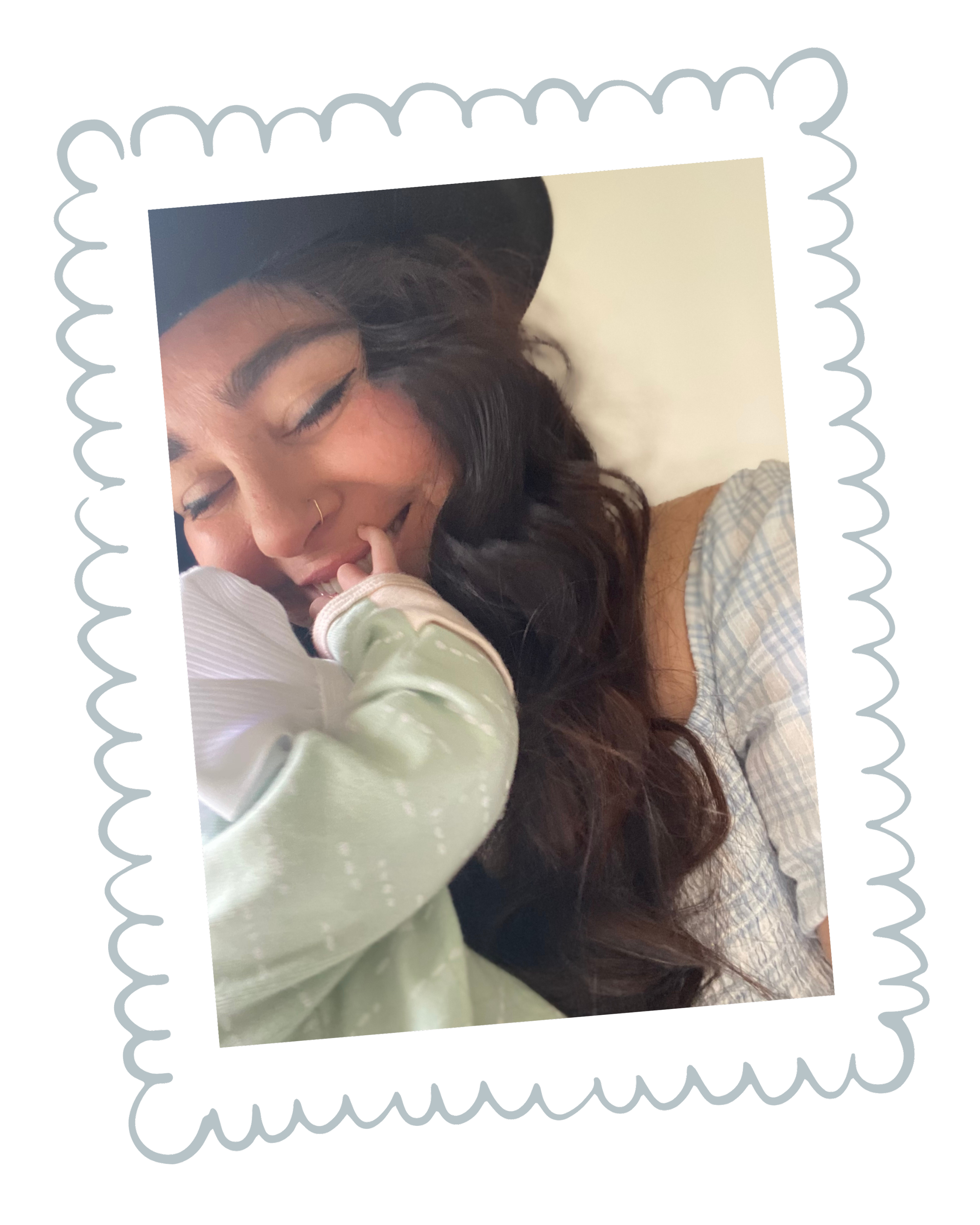 A woman with long dark hair and a nose piercing is smiling with her eyes closed, holding a hand near her face. She is lying on a pillow and wearing a light-colored, plaid shirt.
