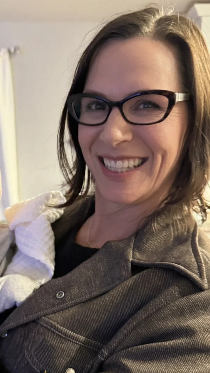 A woman with shoulder-length brown hair, glasses, and a broad smile in an indoor setting with a plain wall and curtains in the background.