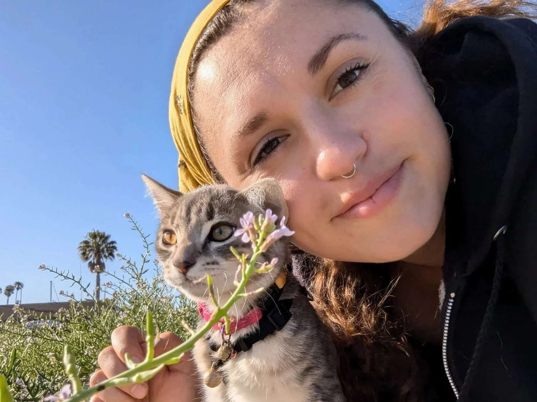 A woman with a septum piercing and a yellow headscarf taking a selfie outdoors with her gray and white cat next to a flowering plant, against a clear blue sky and palm trees in the background.