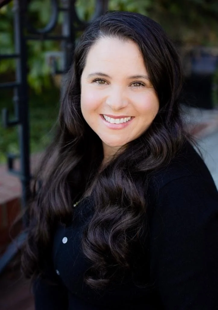 A woman with long dark hair smiling outdoors, standing near a metal garden trellis with greenery in the background.