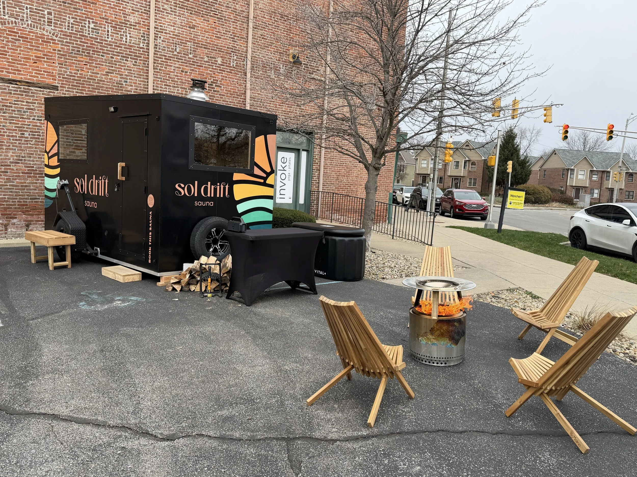 Outdoor seating area with four wooden chairs arranged around a firepit with flames, next to a small black sausage-shaped sauna truck labeled 'sol drift sauna'. The area is on an asphalt surface outside a brick building with leafless trees, parked cars, and traffic lights in the background.