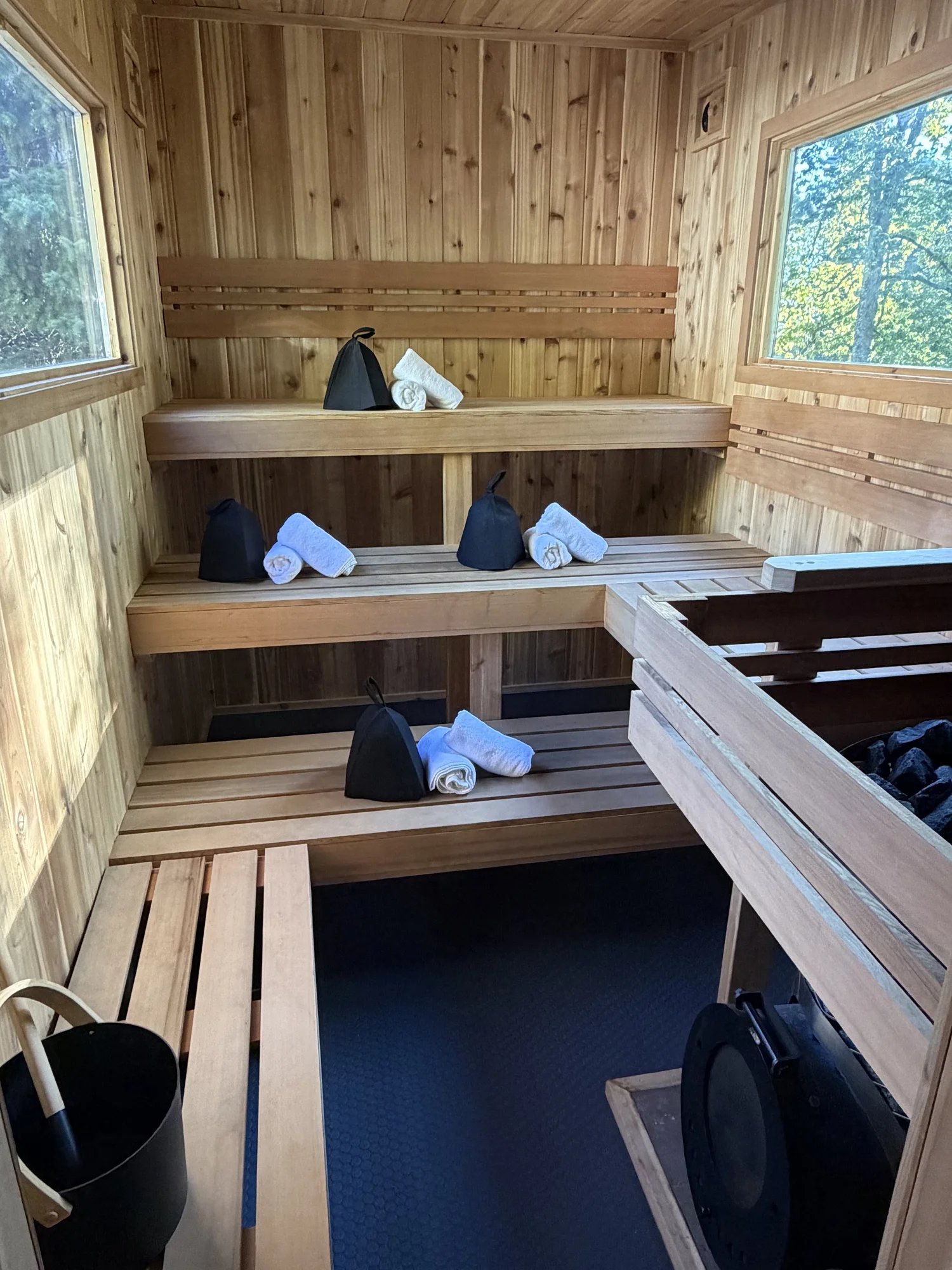 Inside a wooden sauna with tiered benches, towels, and black bags, with windows showing trees outside.