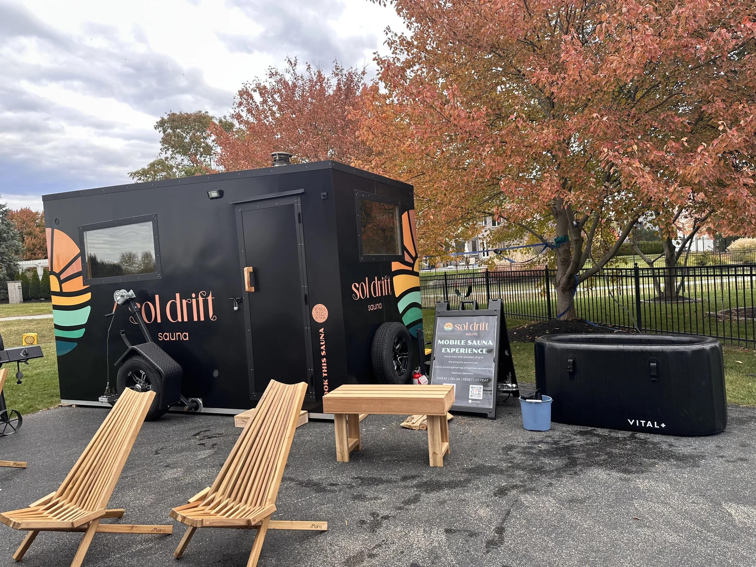 A black mobile sauna trailer with colorful logo and text, outdoor setting with autumn trees, two wooden lounge chairs, a signboard about the mobile sauna experience, a large black speaker, and a blue bucket.
