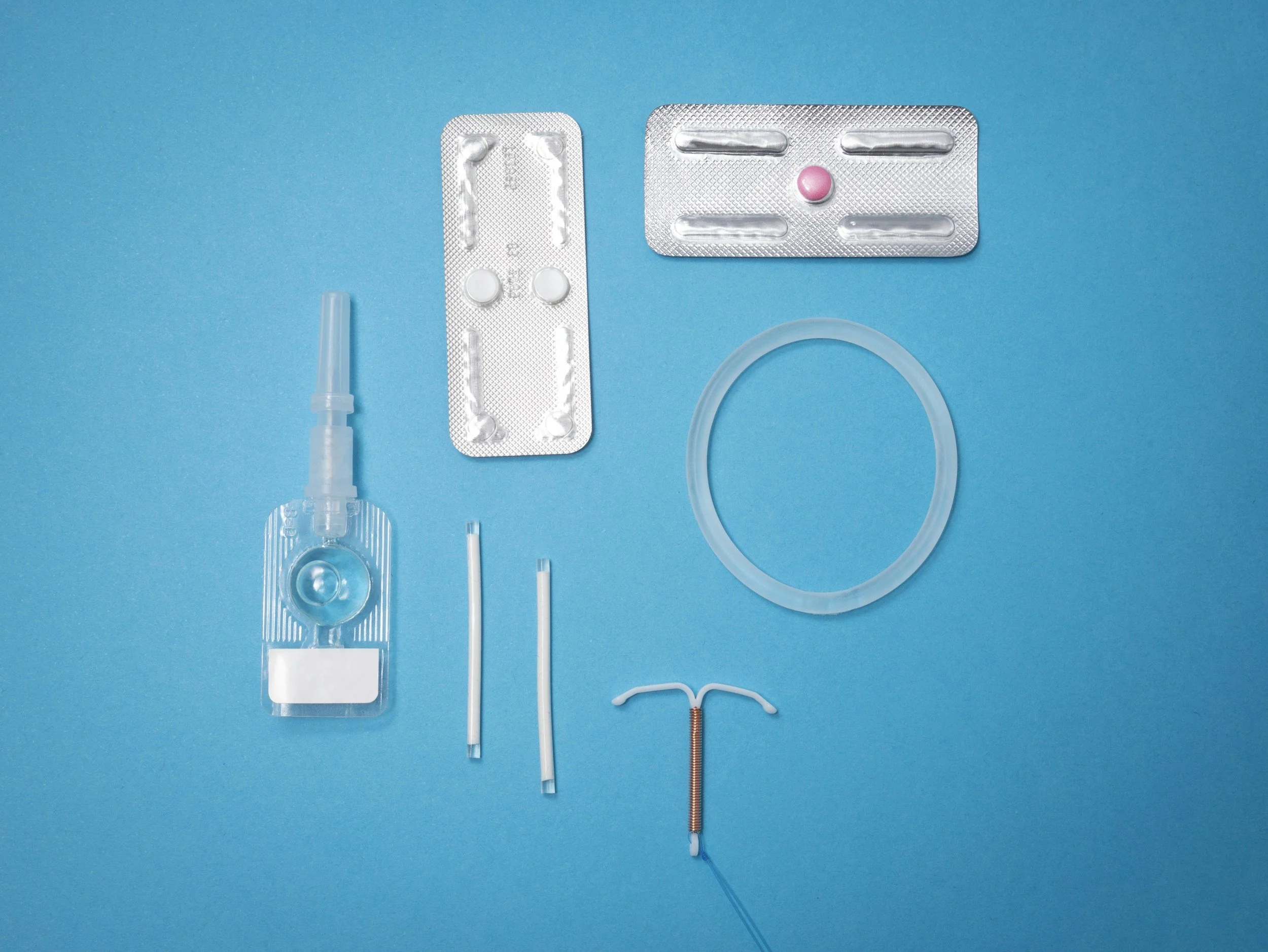 Various medical devices and supplies on a blue background, including a nasal spray bottle, blister packs with pills, a silicone ring, a small needle with a safety cover, and other small medical components.