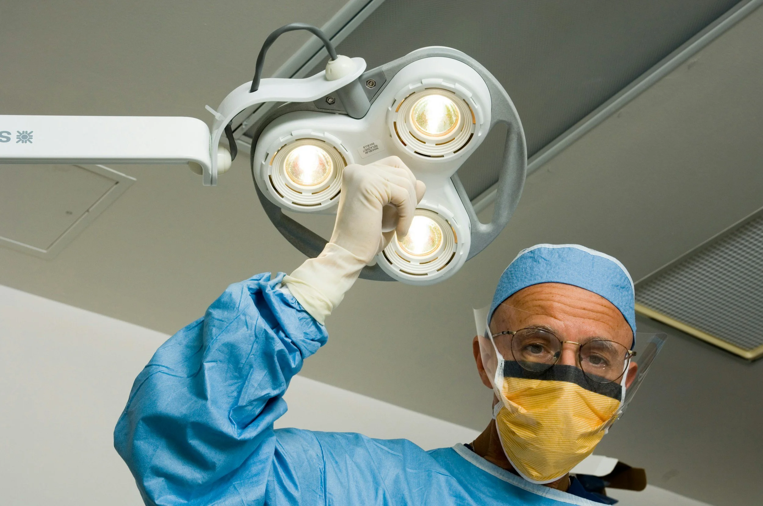 A surgeon in scrubs, wearing a face mask, glasses, and a surgical cap, adjusts a surgical light overhead in an operating room.