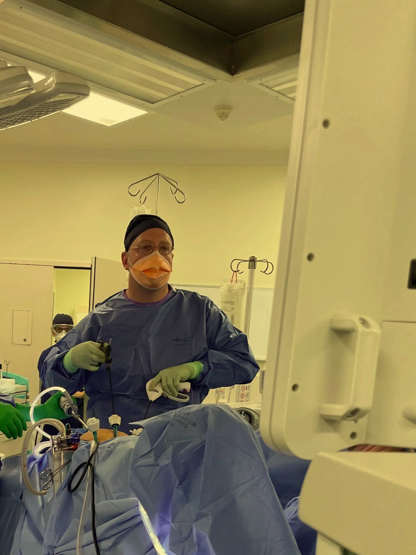 A medical professional wearing scrubs, a face mask, gloves, and glasses is operating in an operating room, assisting with surgery on a patient. hysterectomy, laparoscopy, endometriosis, pelvic pain, period pain, abnormal bleeding.
