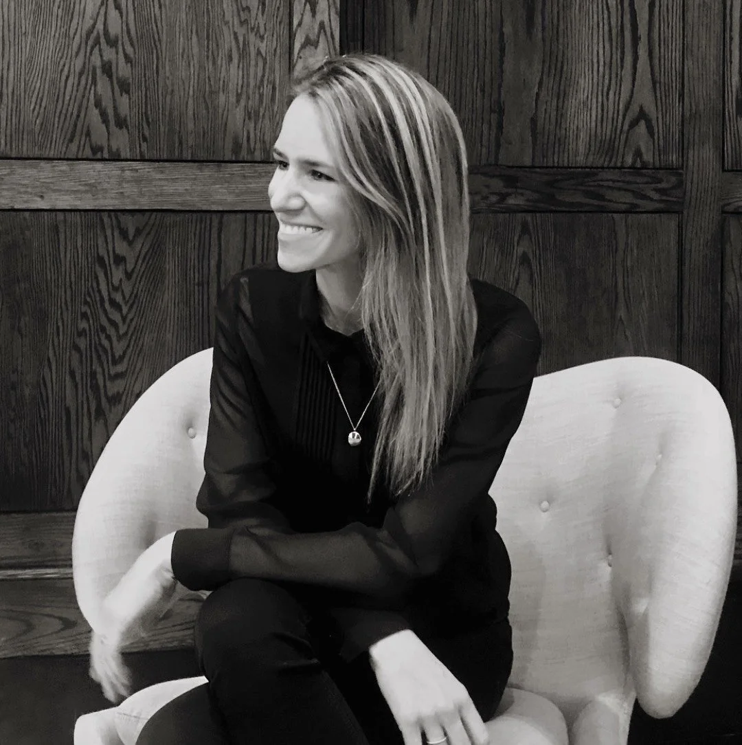 A woman with long hair and a necklace sitting on a light-colored chair, smiling and looking to the side against a dark wooden wall.