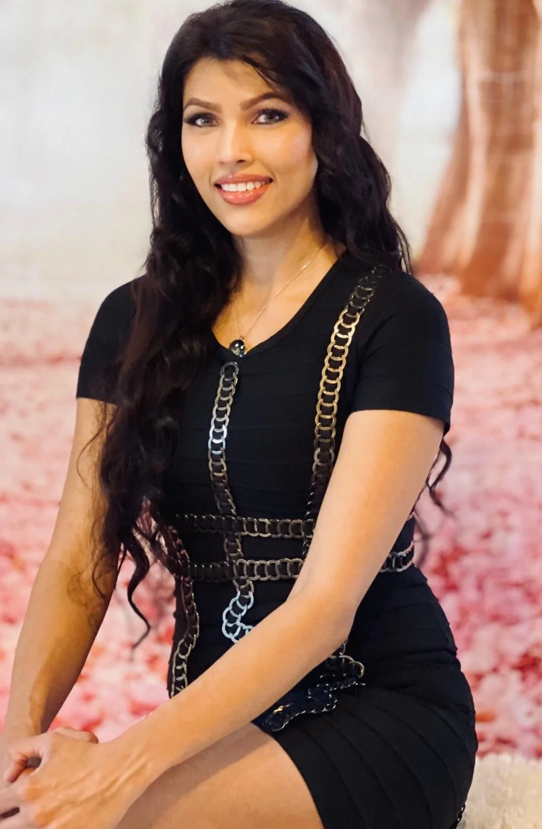 A woman with long dark wavy hair wearing a black dress with metallic chain accessories, smiling and sitting indoors with a pinkish background.