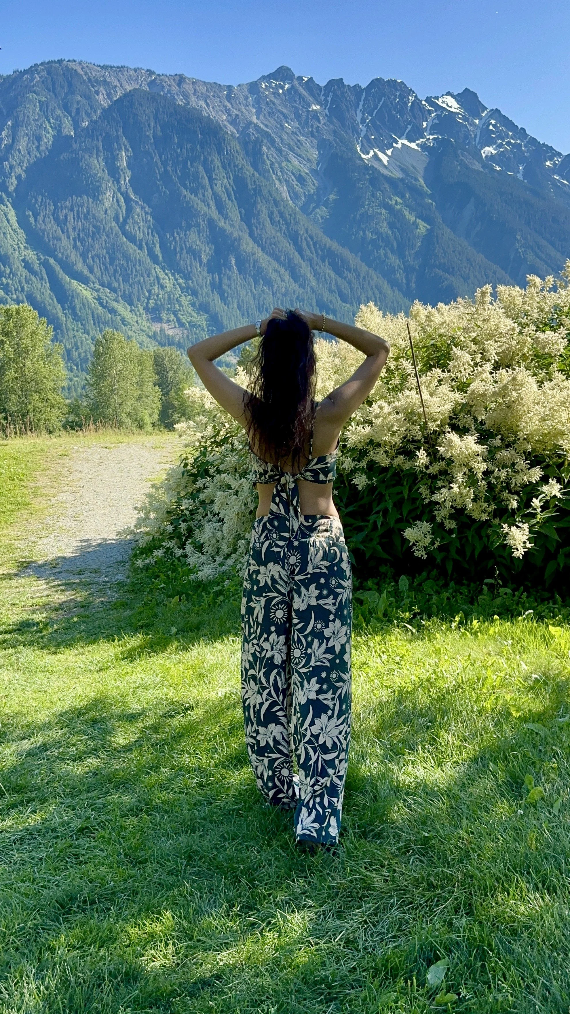 Woman standing on a grassy path with her back to the camera, wearing a floral outfit, surrounded by lush greenery and white flowers, with tall mountains in the background under a clear blue sky.
