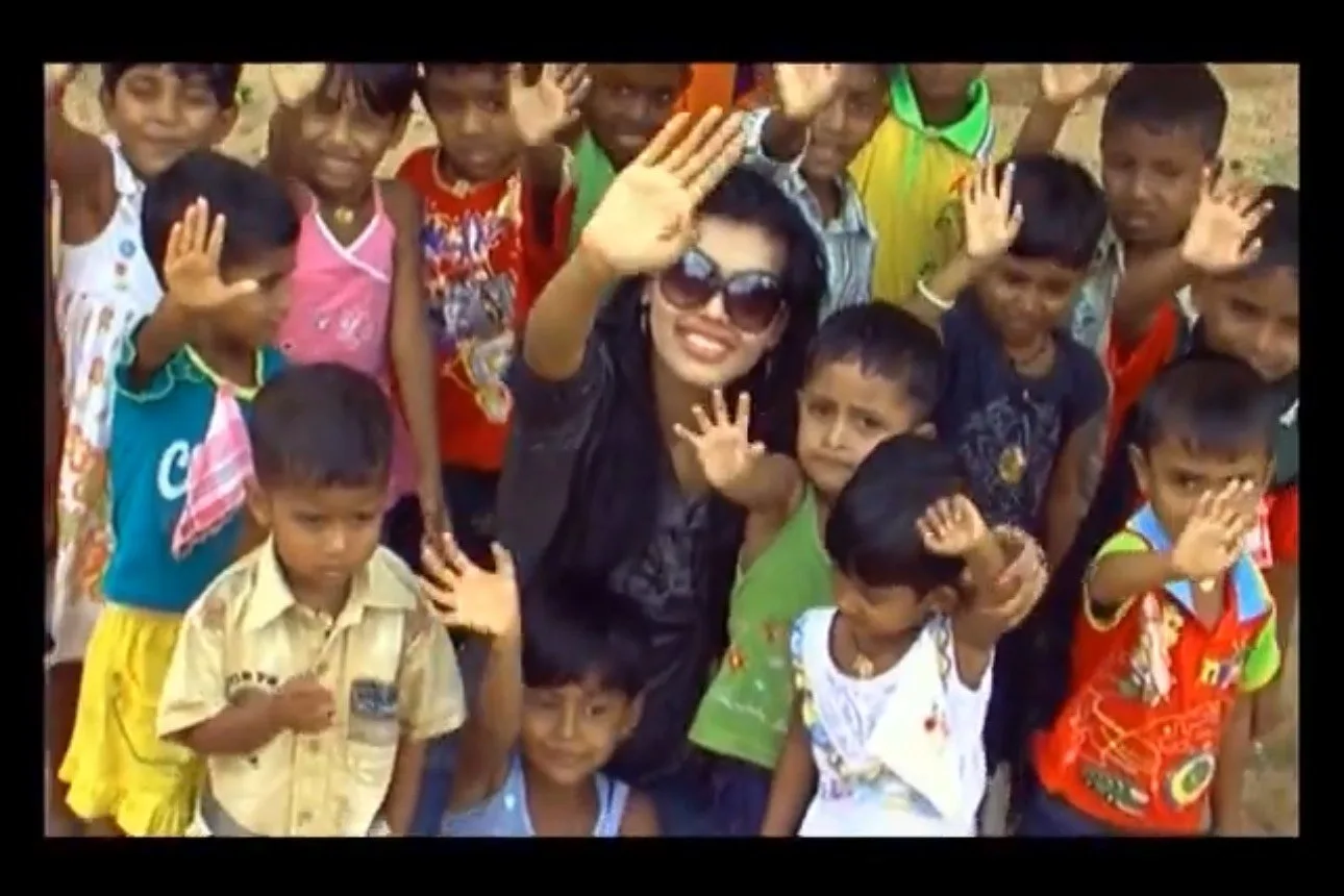 A woman in sunglasses waving with children around her, some waving and some covering their eyes, outdoors.