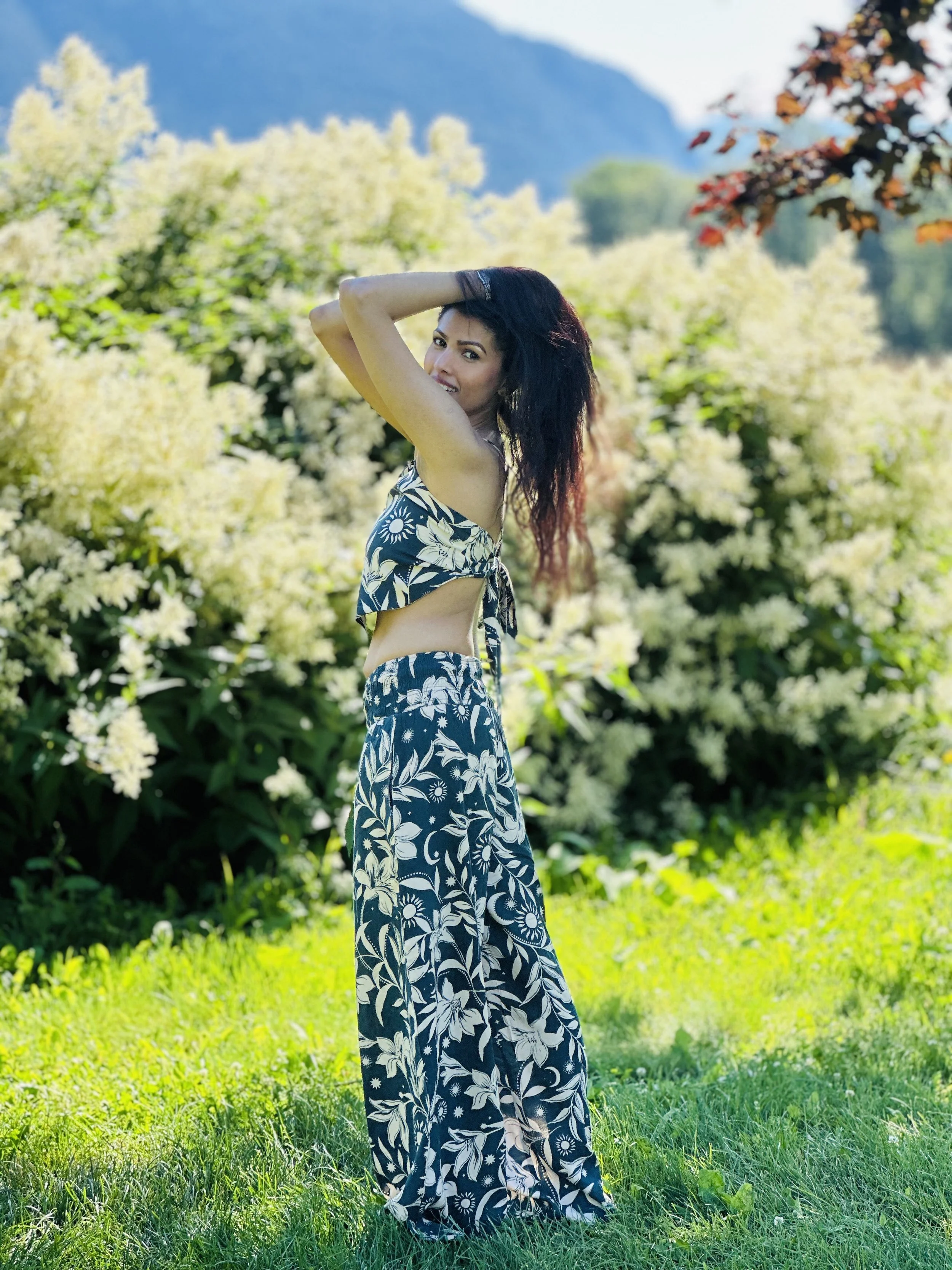 A woman in a black and white floral outfit poses outdoors with her left arm raised, standing on green grass, with white flowering bushes and trees in the background on a sunny day.