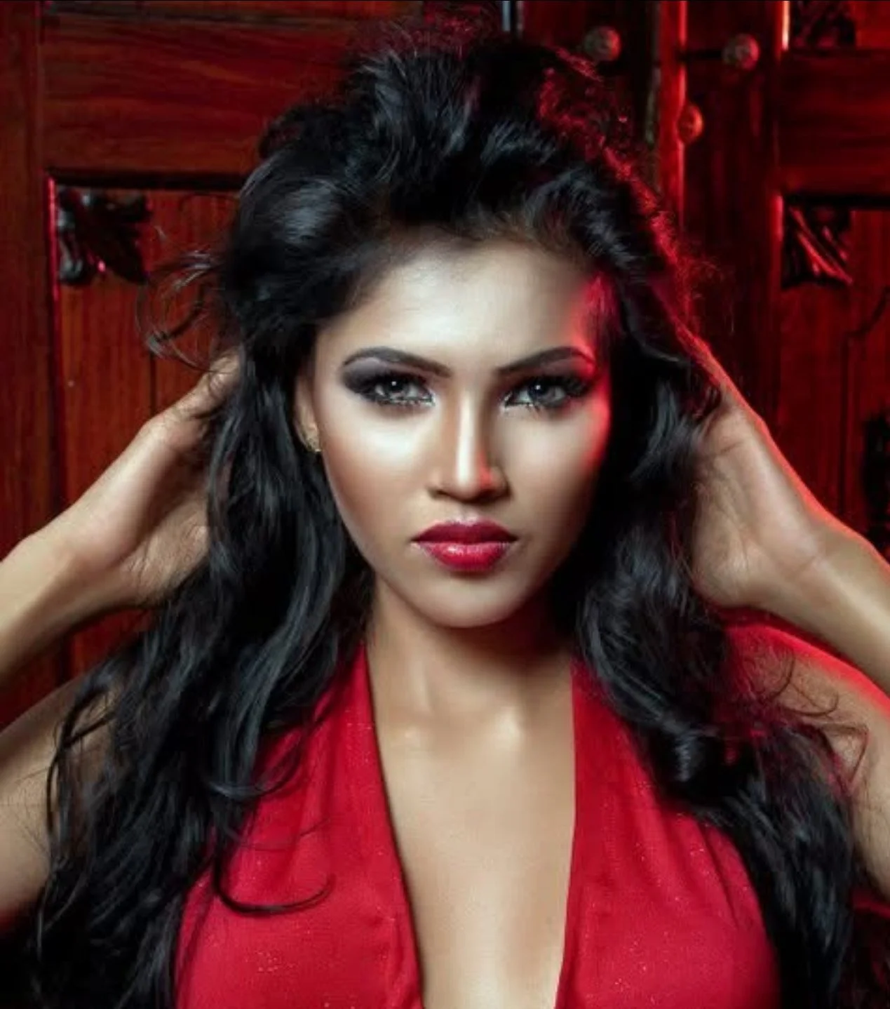 A woman with long dark hair, makeup, and red lipstick poses in front of a wooden background.