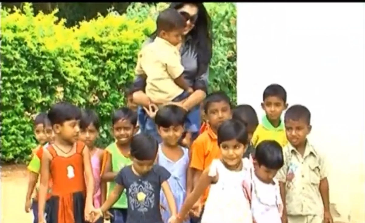A woman holds a young boy in her arms with a group of children standing in front of her outside, in front of a yellow-green hedge and a white wall, during daytime.