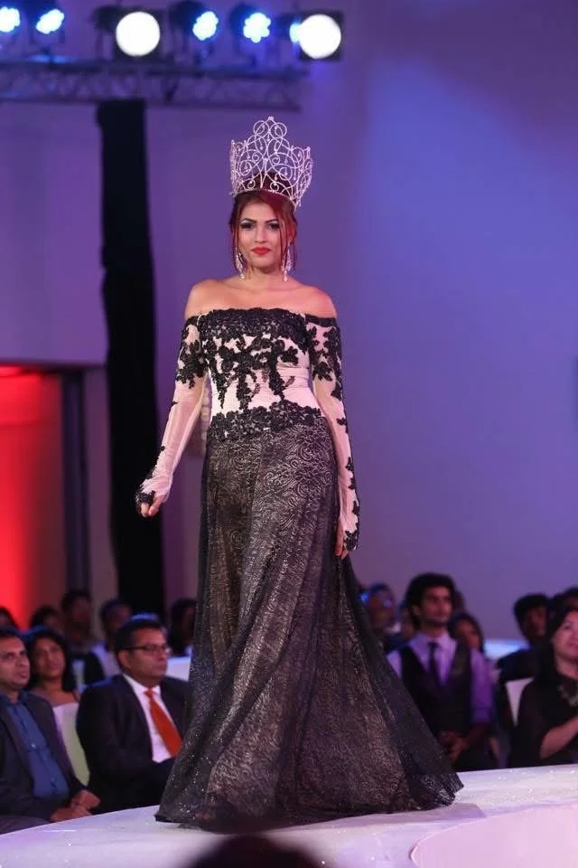 A woman in a glamorous black and pink gown with lace detailing walks on a runway during a fashion show. She wears a large, ornate crown and has long earrings, with an audience watching in the background.