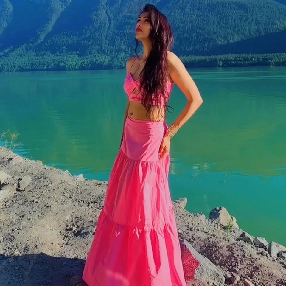 Woman in a pink dress posing by a lake with mountain ranges in the background.