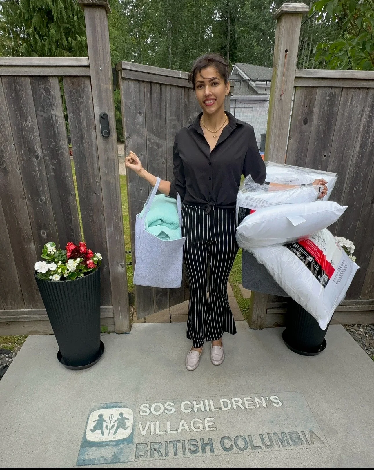 A woman standing outside in front of a wooden fence, holding shopping bags with clothing and pillows, on a concrete sidewalk that reads 'SOS CHILDREN'S VILLAGE BRITISH COLUMBIA.' She is making a playful face, wearing a black button-up shirt, black an