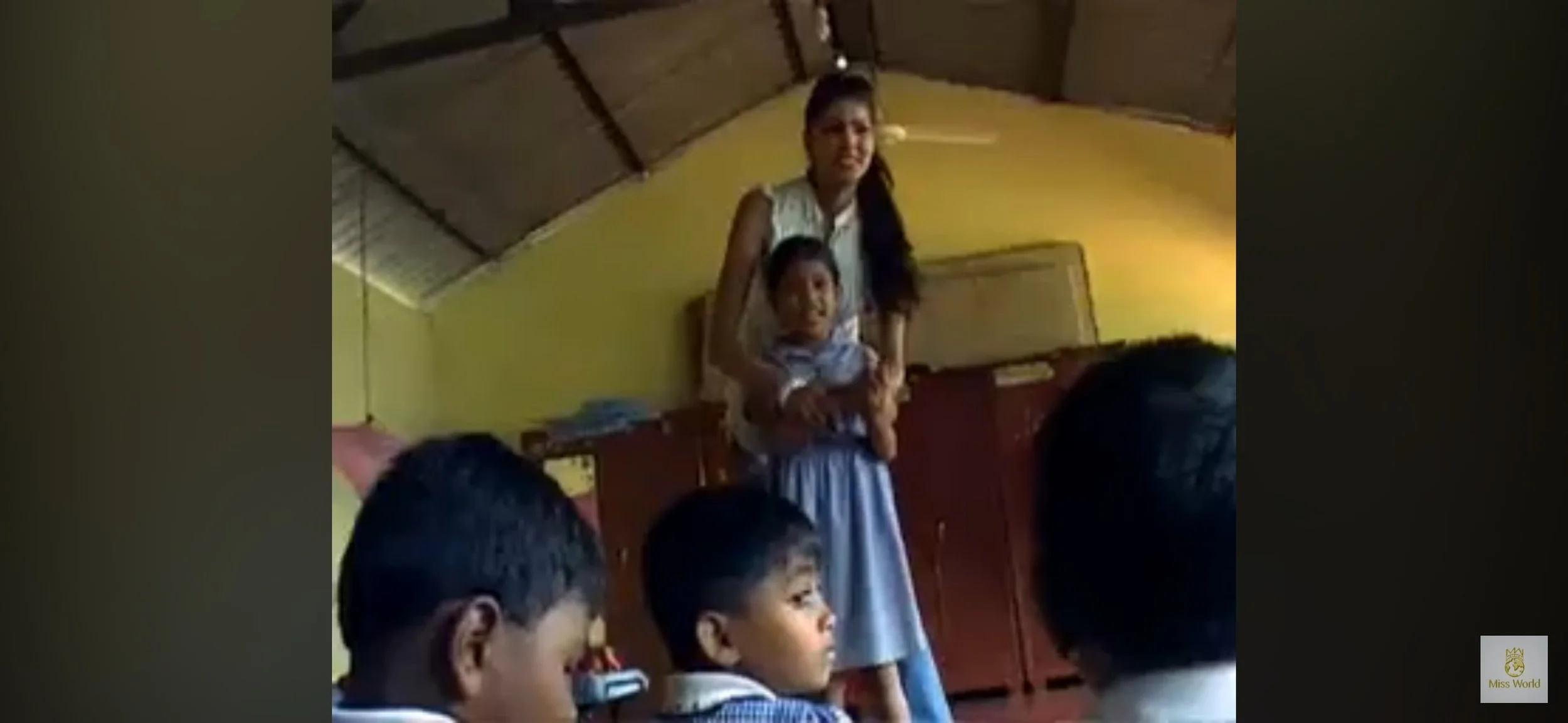 A woman standing in a classroom holding a young boy, with three other children sitting in the foreground. The classroom has yellow walls and a wooden cabinet.