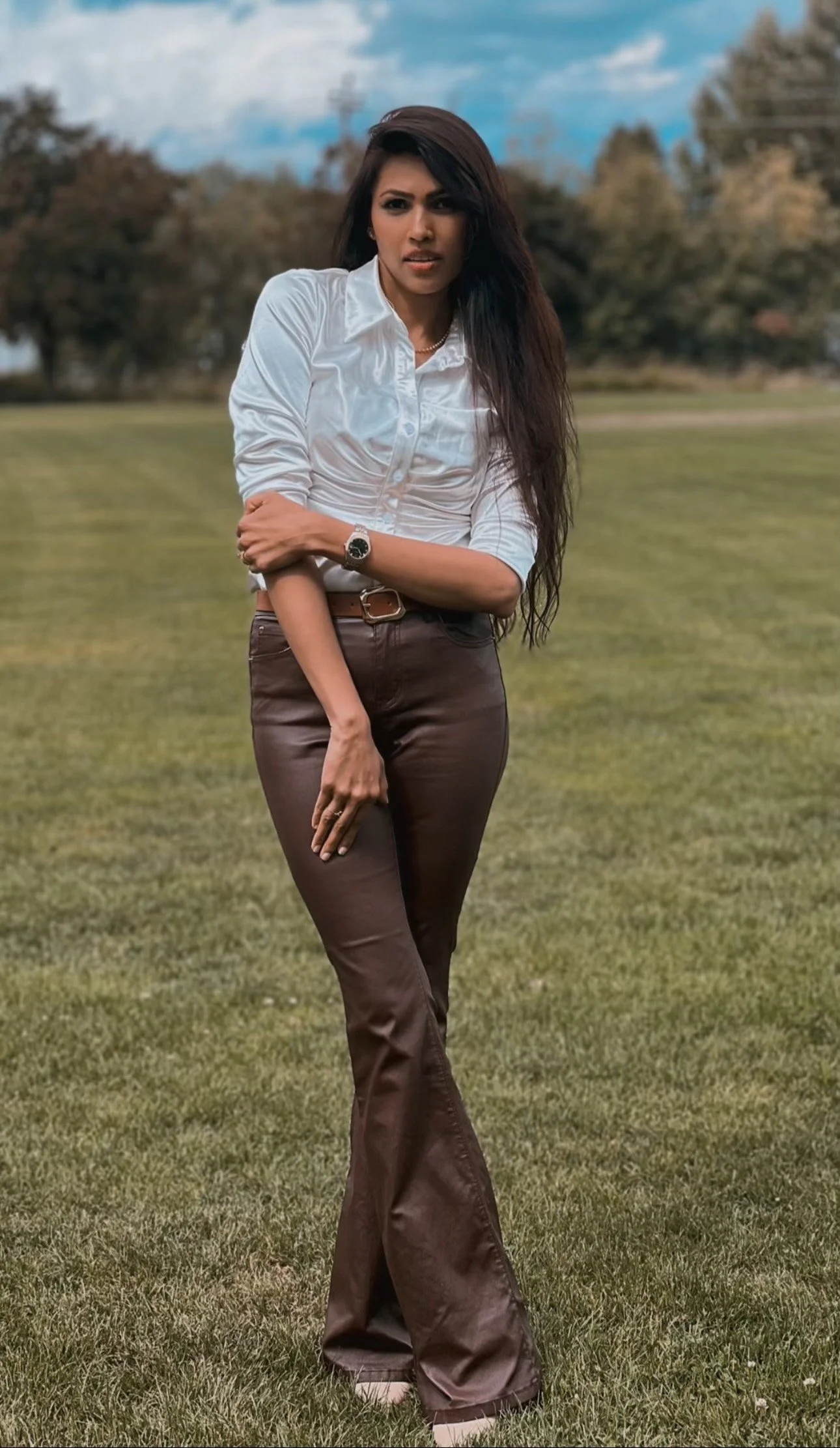 A woman standing on a grassy field, dressed in a white blouse and brown pants, with trees and a partly cloudy sky in the background.